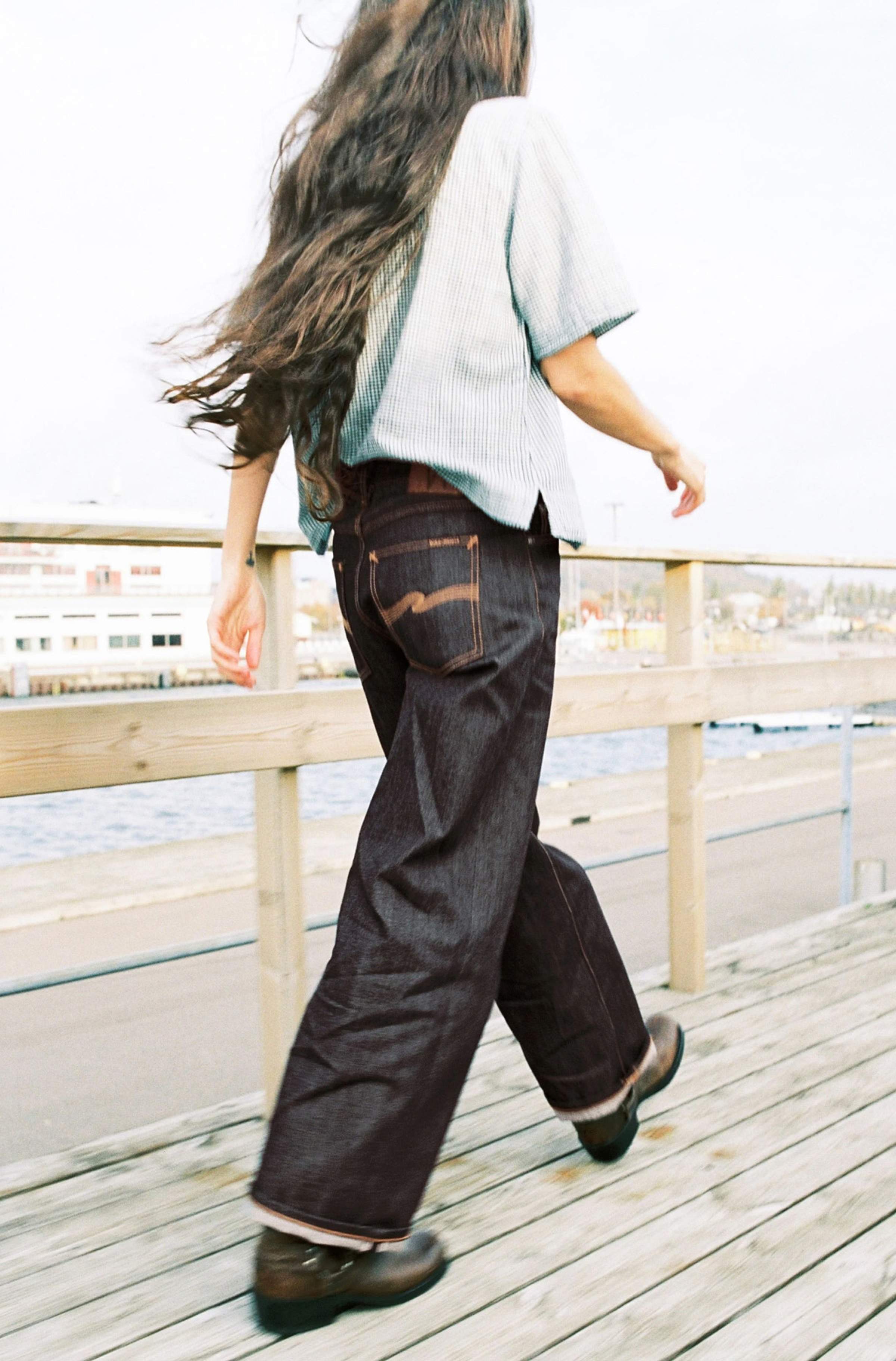 Model walking on a wooden pier in dark raw selvedge Nudie Jeans with a striped short-sleeve top.