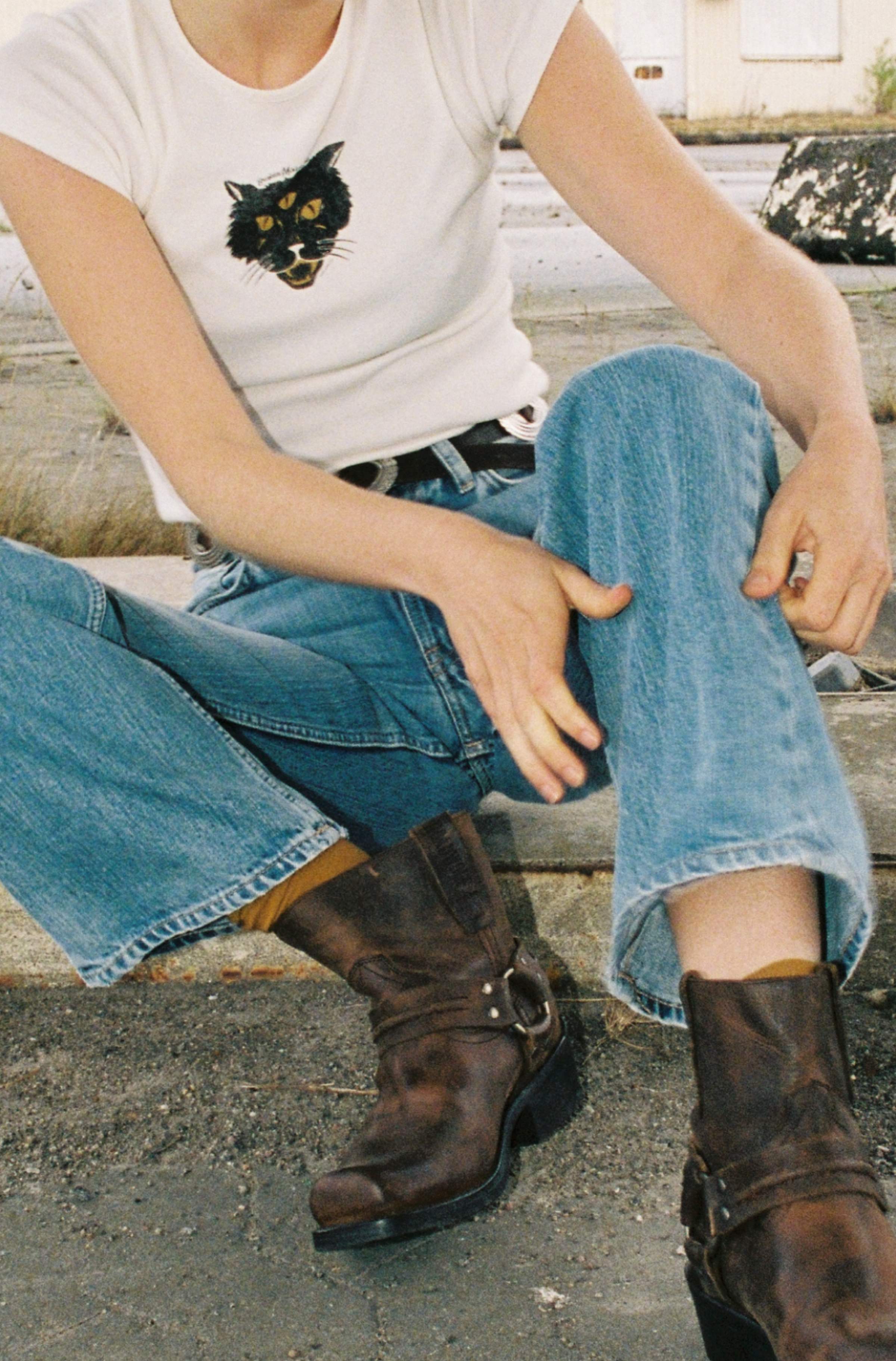 Model in white cat graphic tee and Nudie Jeans washed blue denim with brown harness boots, sitting on pavement.