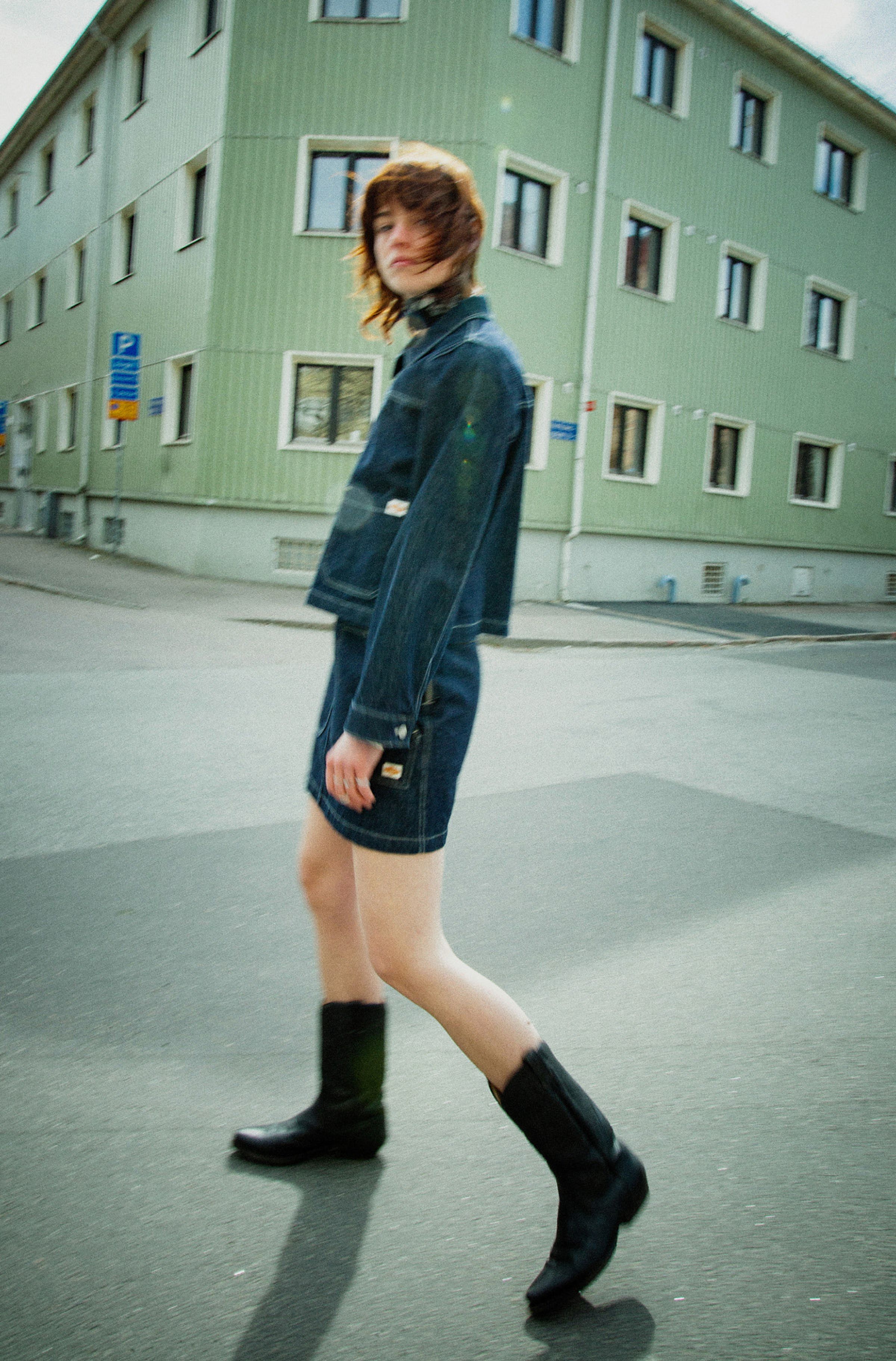 Person in a denim jacket and denim skirt walking on a street near a green building with multiple windows. Blurred motion effect.