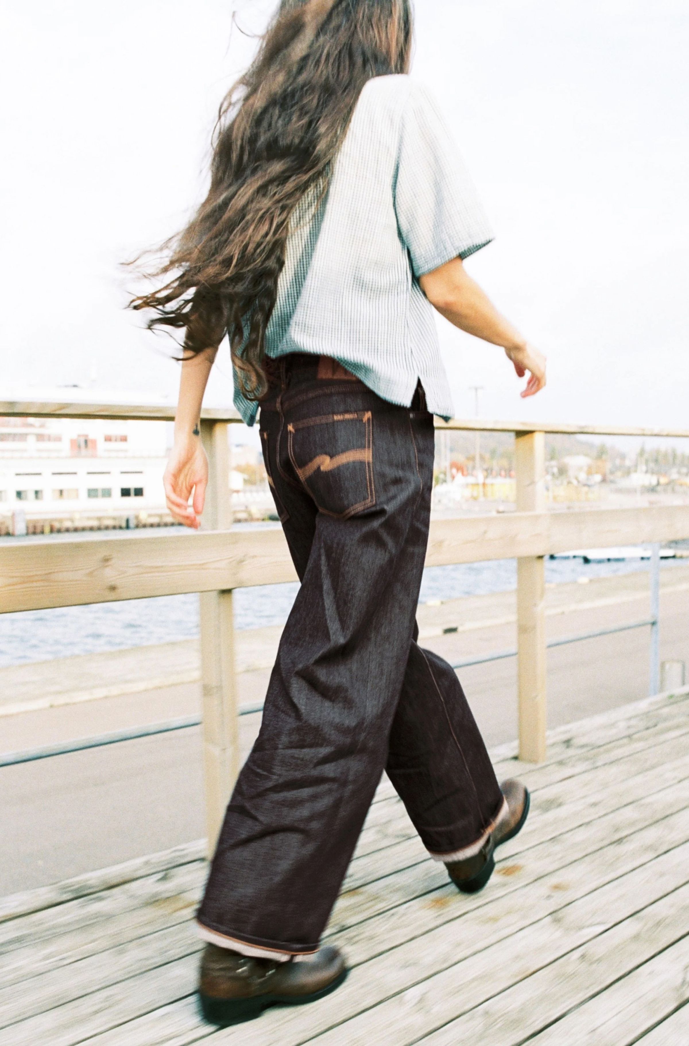 Person with long hair walking on a wooden boardwalk, wearing a striped shirt, dark wide-leg jeans, and boots, with water and buildings in the background.