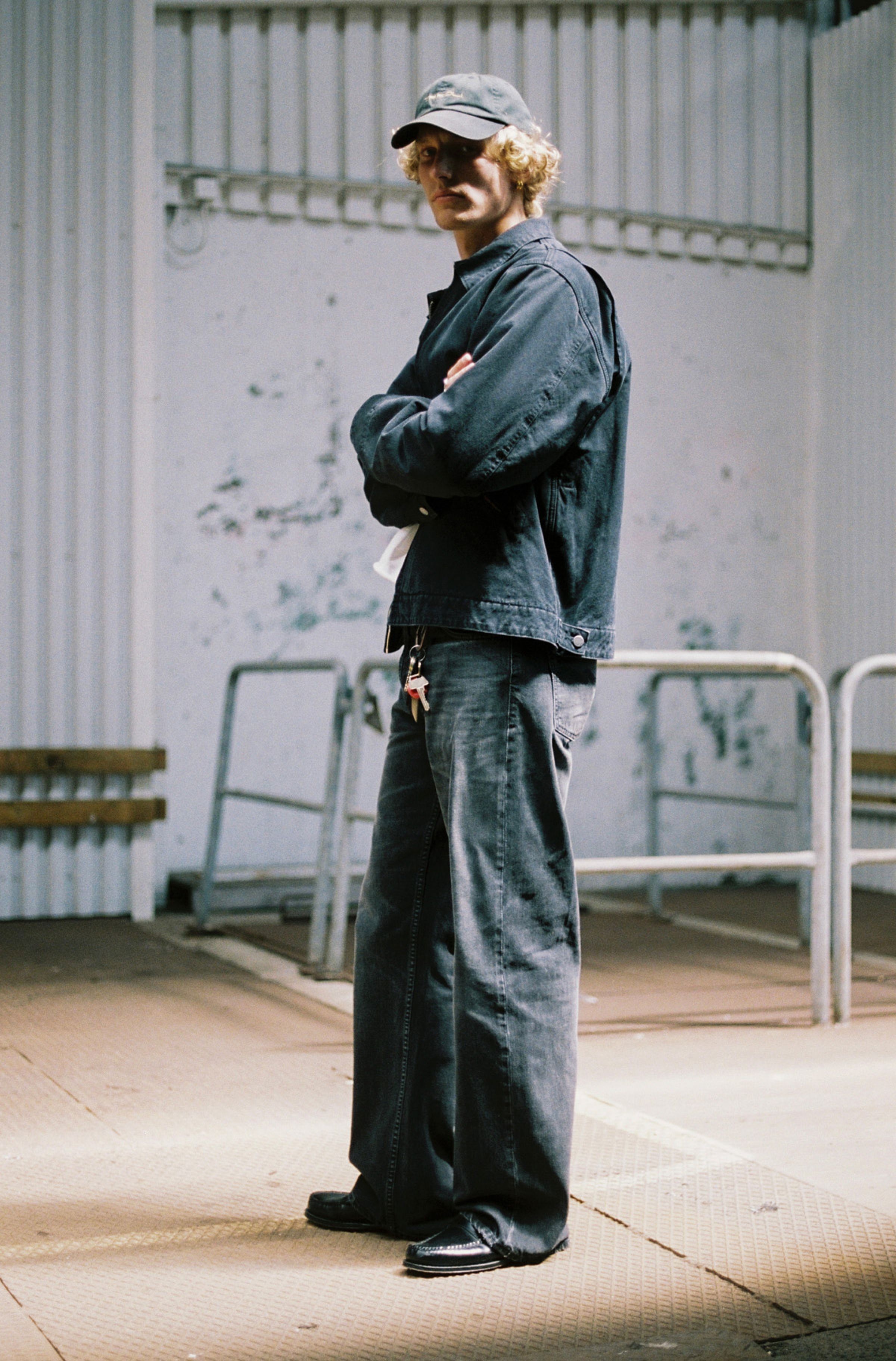 Person in a black jacket and black jeans and a cap stands with arms crossed in an industrial setting with metal railings and a textured wall.