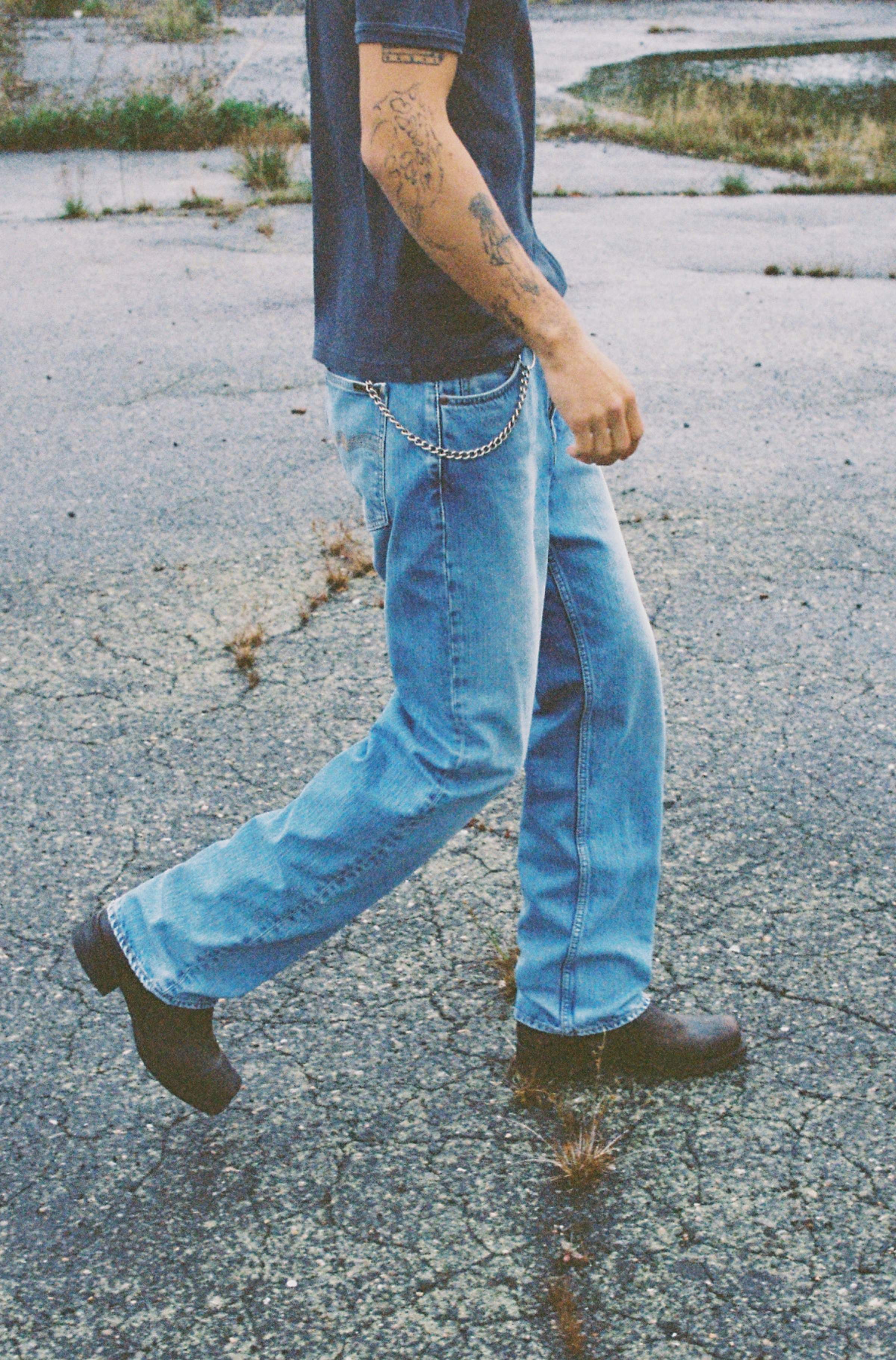 Model walking on cracked pavement in light wash Nudie Jeans prewashed blue jeans with wallet chain and brown boots.