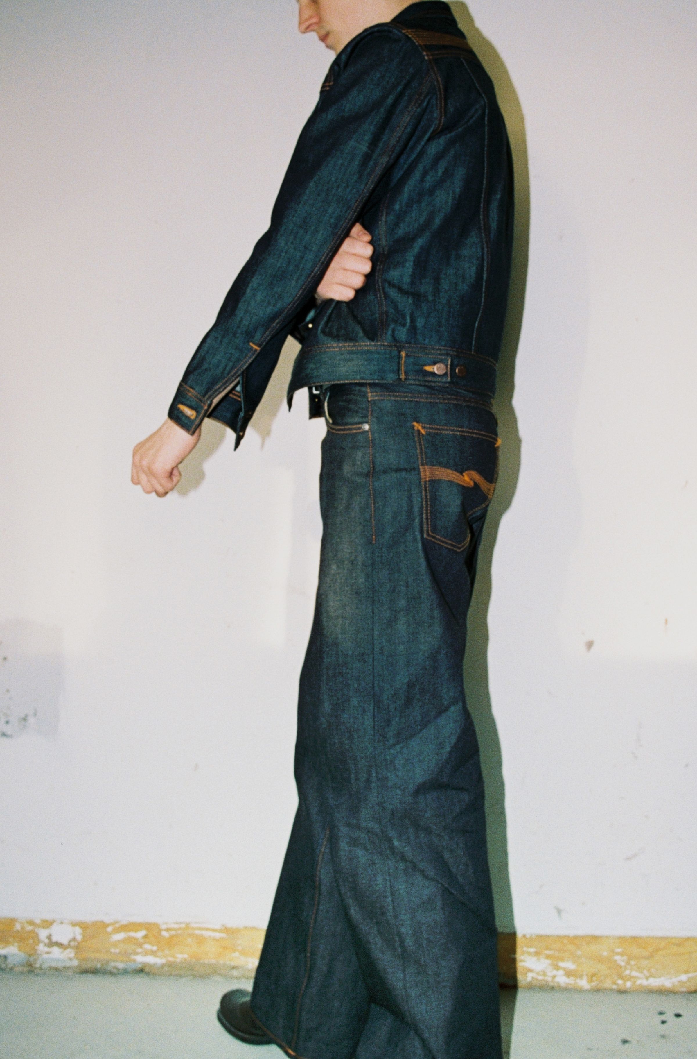 Person wearing a dry raw denim jacket and wide-leg jeans, standing against a plain wall with a yellow baseboard.