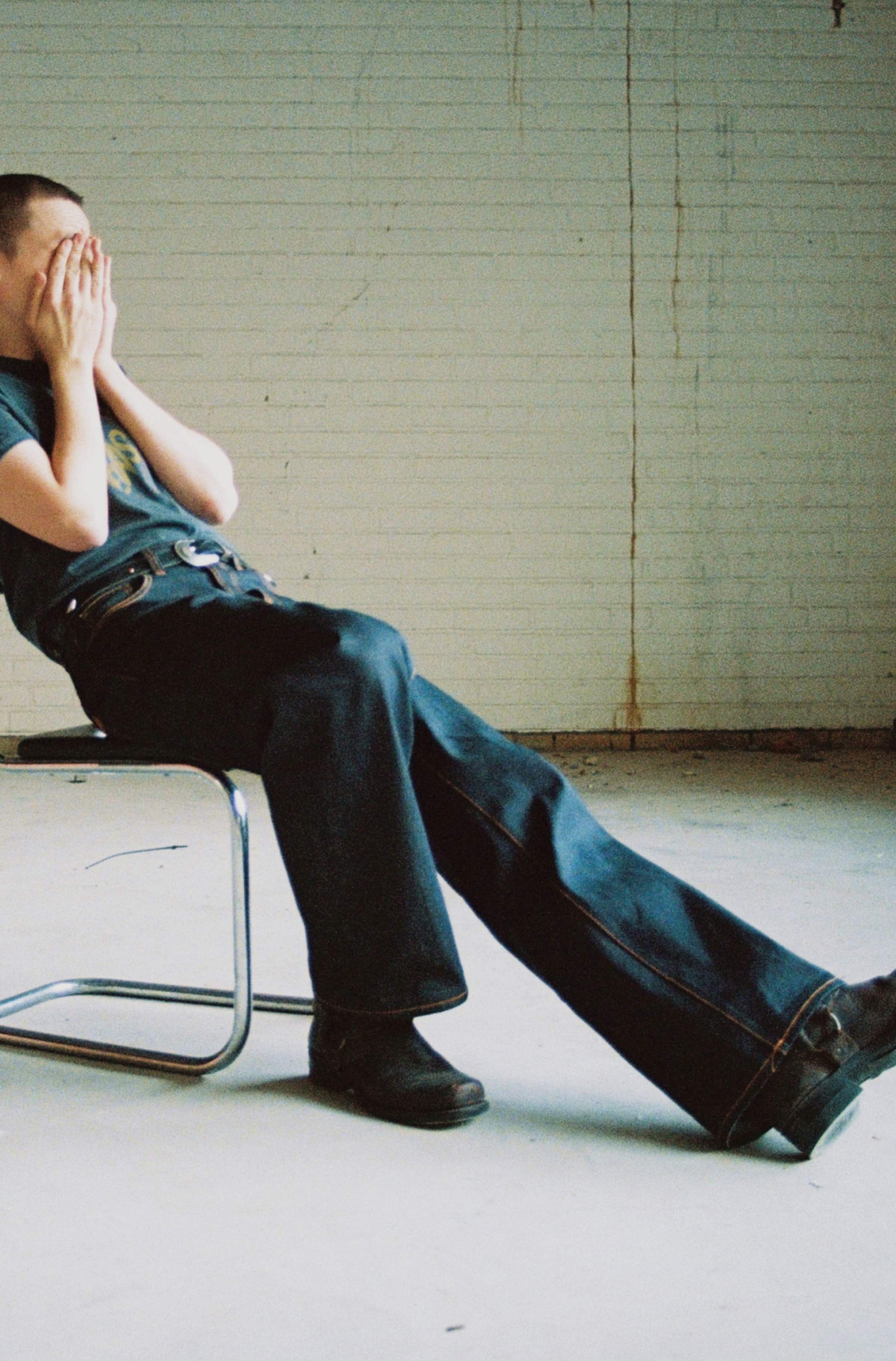 Model in dark raw selvedge Nudie Jeans seated on a tubular chair with hands covering face, legs stretched out.