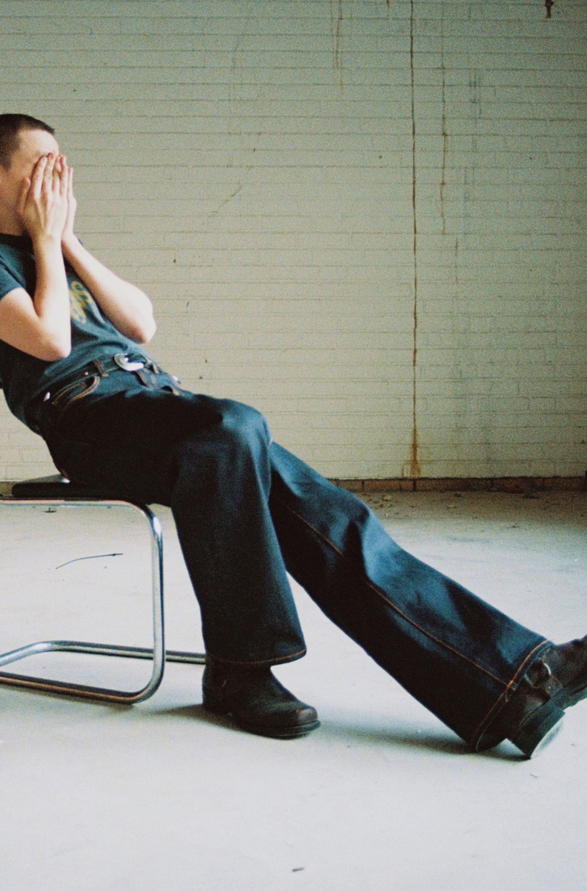 Person sitting on a chair, wearing dry raw jeans and shoes, covering their face with hands, against a white brick wall background.