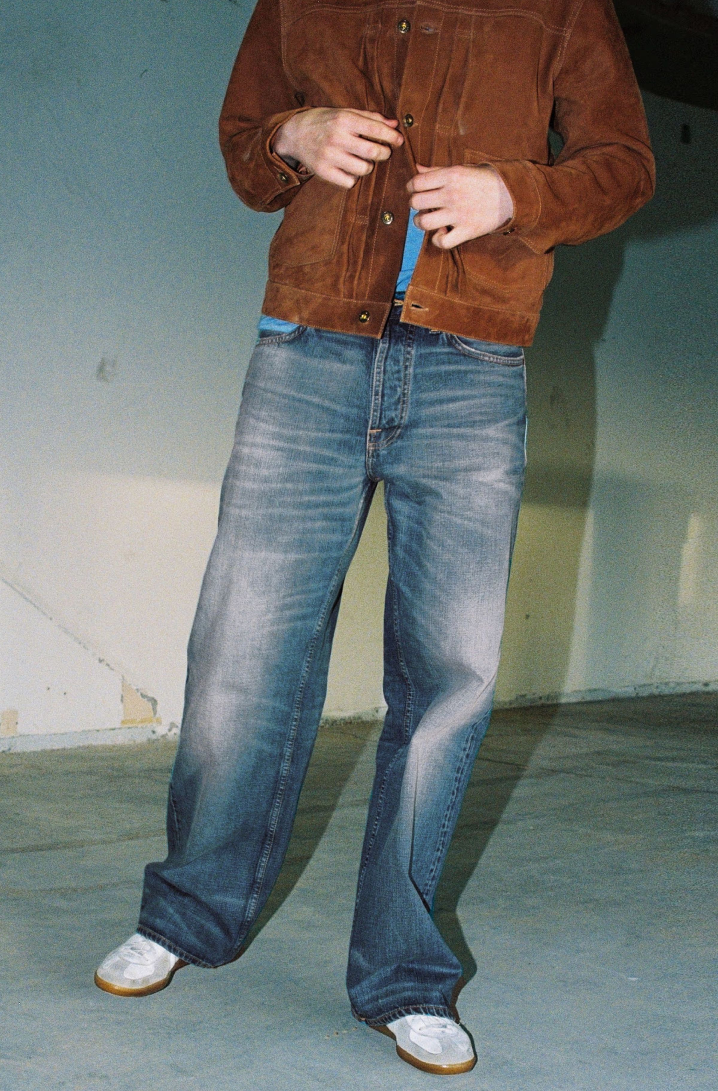 Person in a brown jacket and faded jeans stands in a neutral space, hands adjusting the jacket.