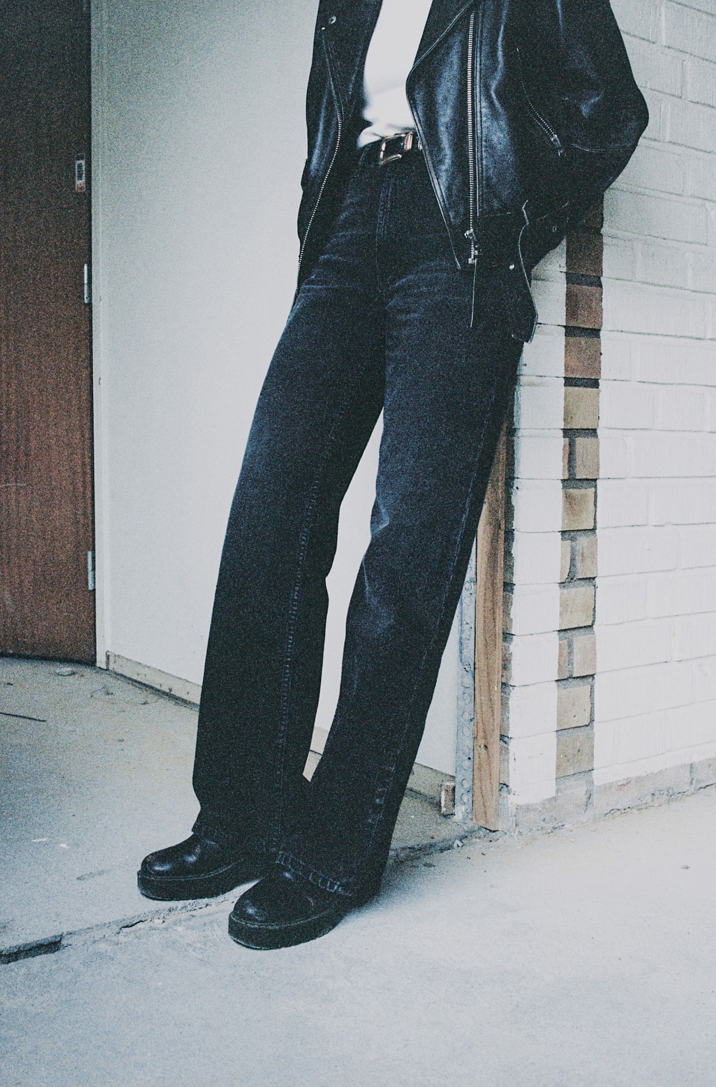 Person in a black leather jacket and black wide-leg jeans leans against a brick wall, with hands in pockets.