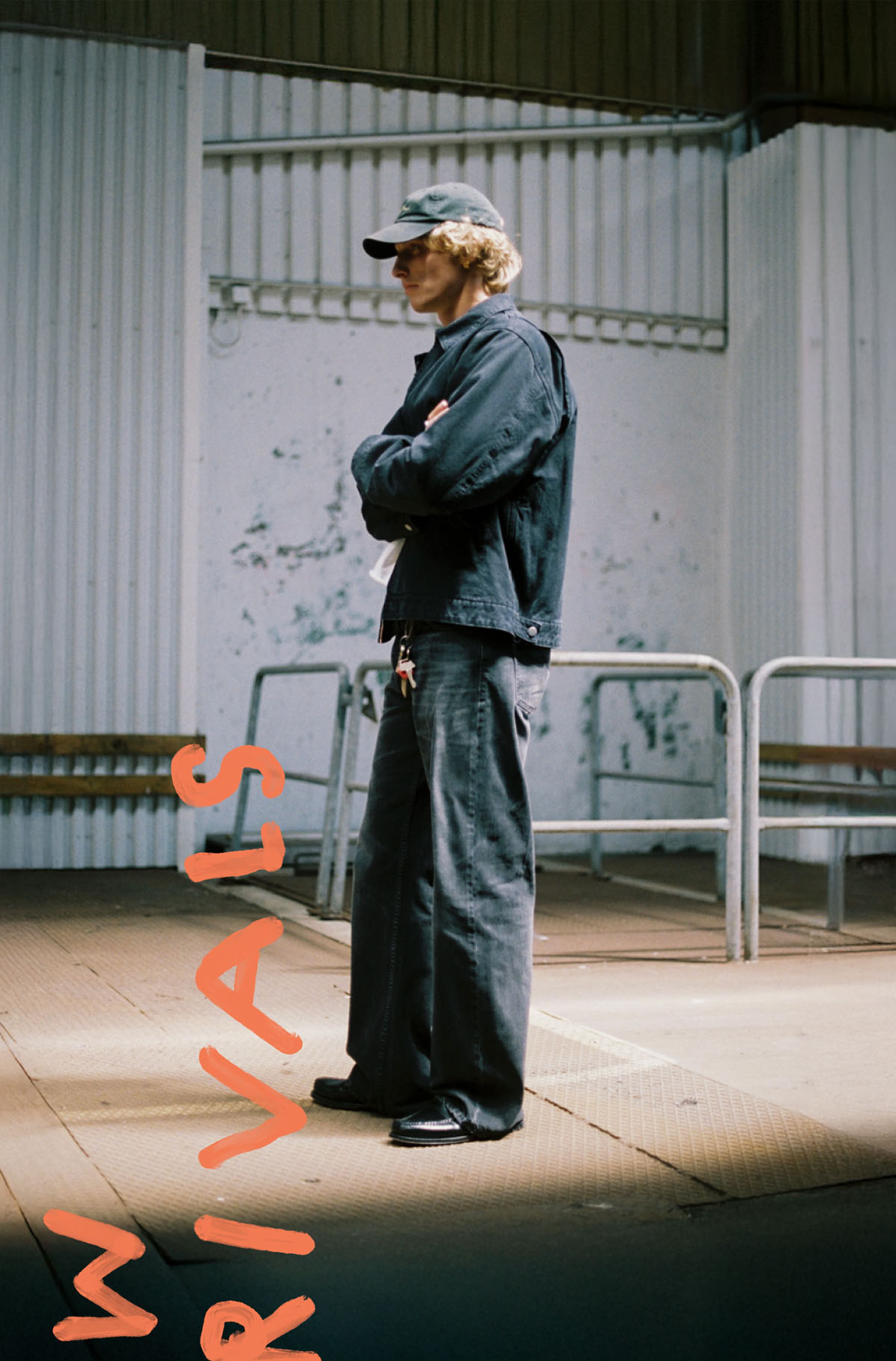 Person in black jacket and pants, wearing a cap, stands with arms crossed. Background features industrial-style metal railings and walls.