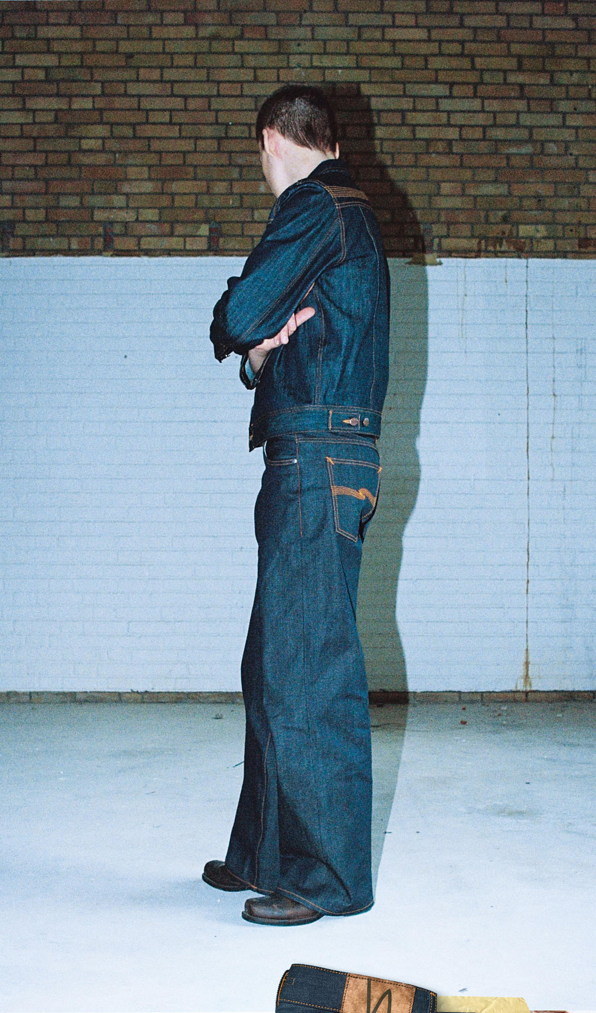 Person in dry, raw denim jacket and wide-leg jeans, standing with arms crossed, facing forward and sideways against a brick and white wall.