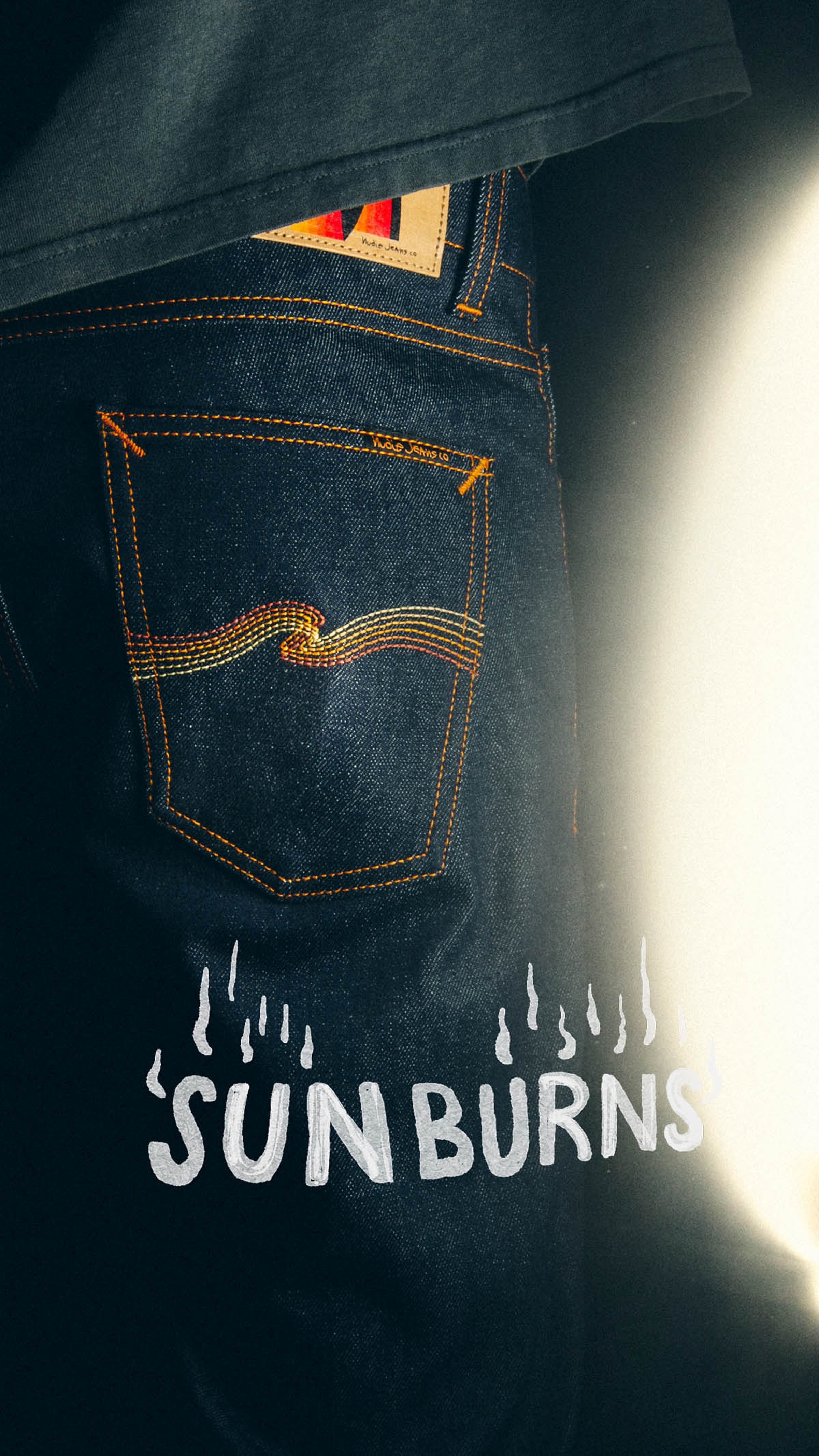 Close-up of denim jeans with orange stitching, featuring the word "SUNBURNS" and flame illustrations on the back pocket.