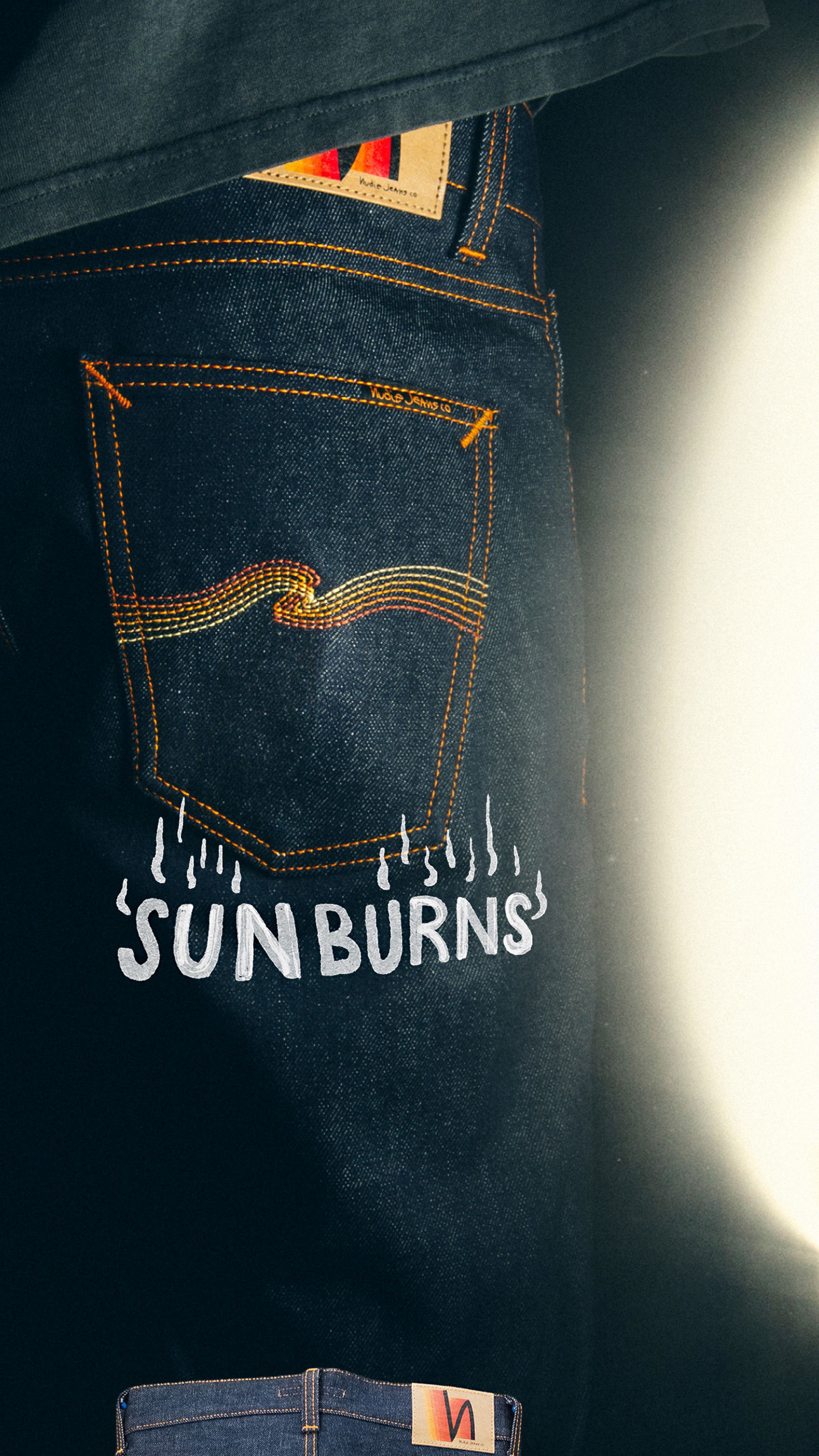 Close-up of denim jeans with "SUN BURNS" printed under the back pocket, highlighted by bright lighting on the right.