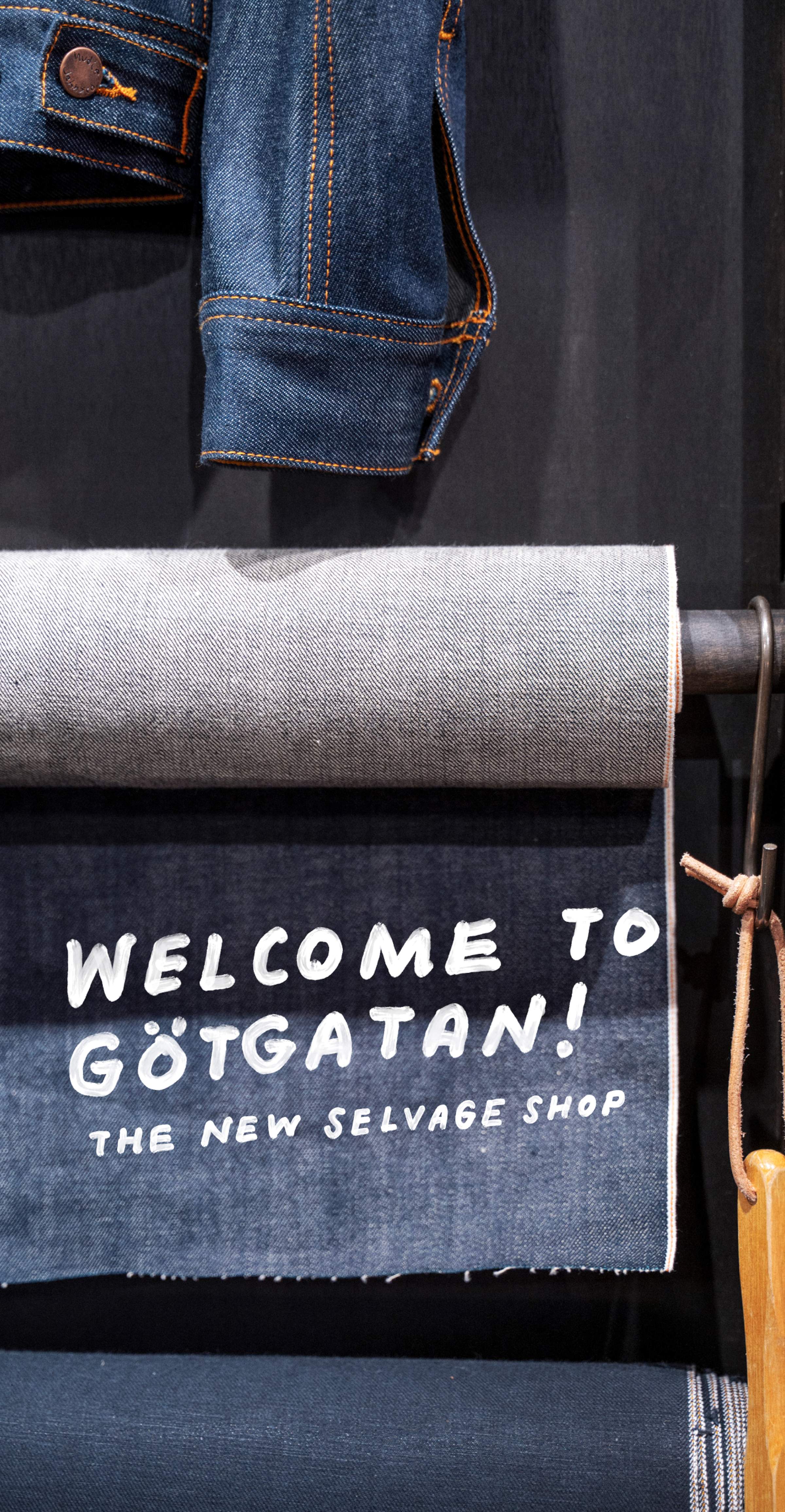 Denim fabric rolls displayed with a sign reading "Welcome to Götgatan! The New Selvage Shop." A denim jacket is in the background.