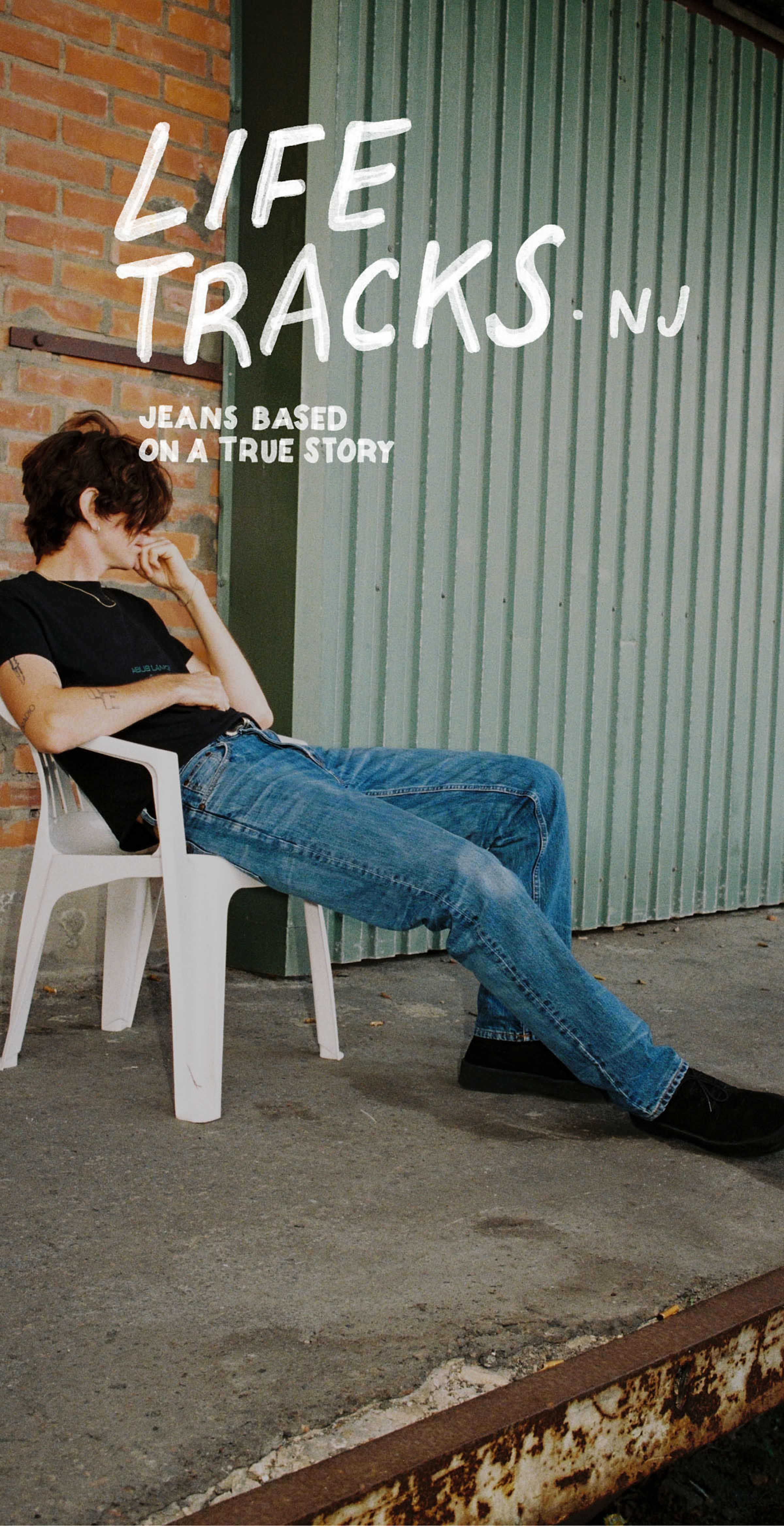 Person in a black shirt and jeans sits in a white chair against a brick and metal wall. Text reads "Life Tracks NJ, Jeans Based on a True Story."