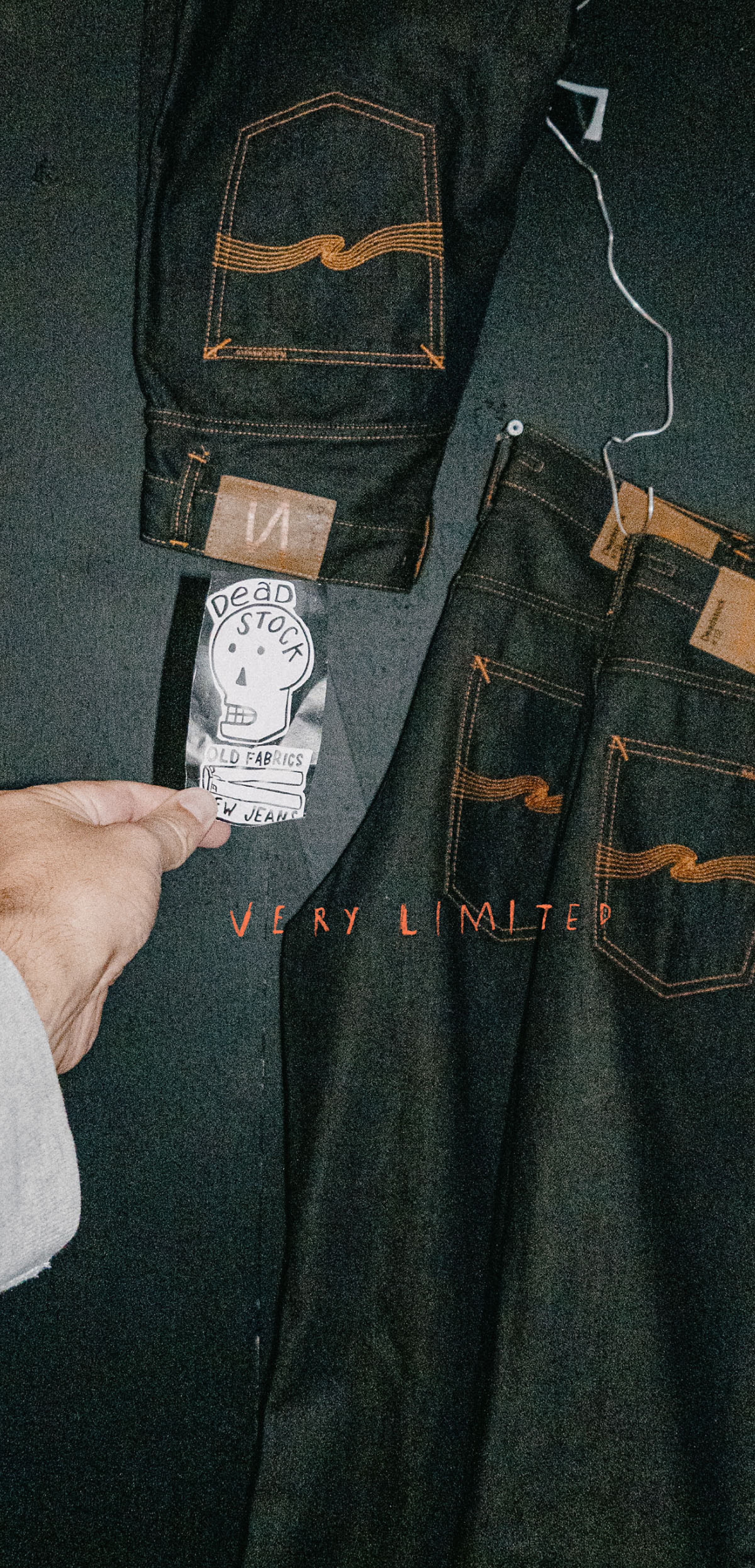 A hand holds a "Deadstock" tag in front of two pairs of dark denim jeans hanging on a black wall, with "Very Limited" written in red.