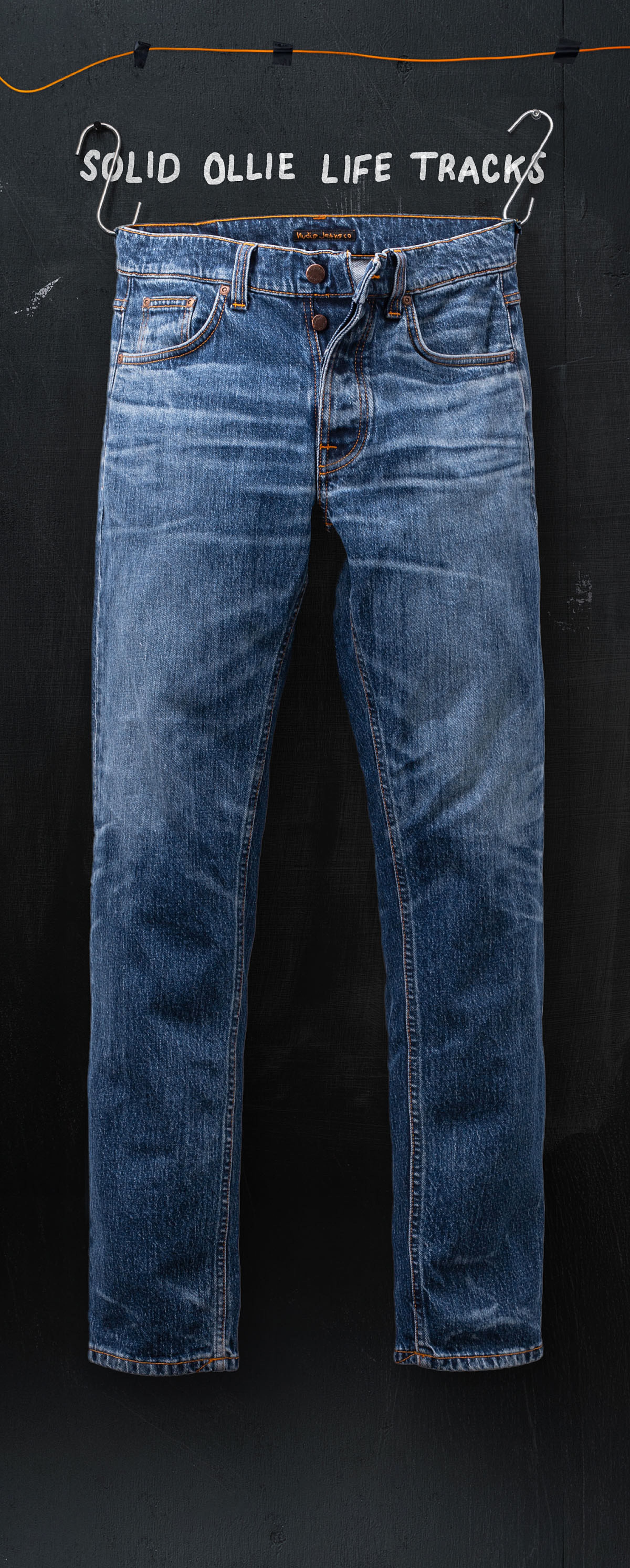 Blue denim jeans hanging against a dark background, with "Solid Ollie Life Tracks" written above on a hanger.