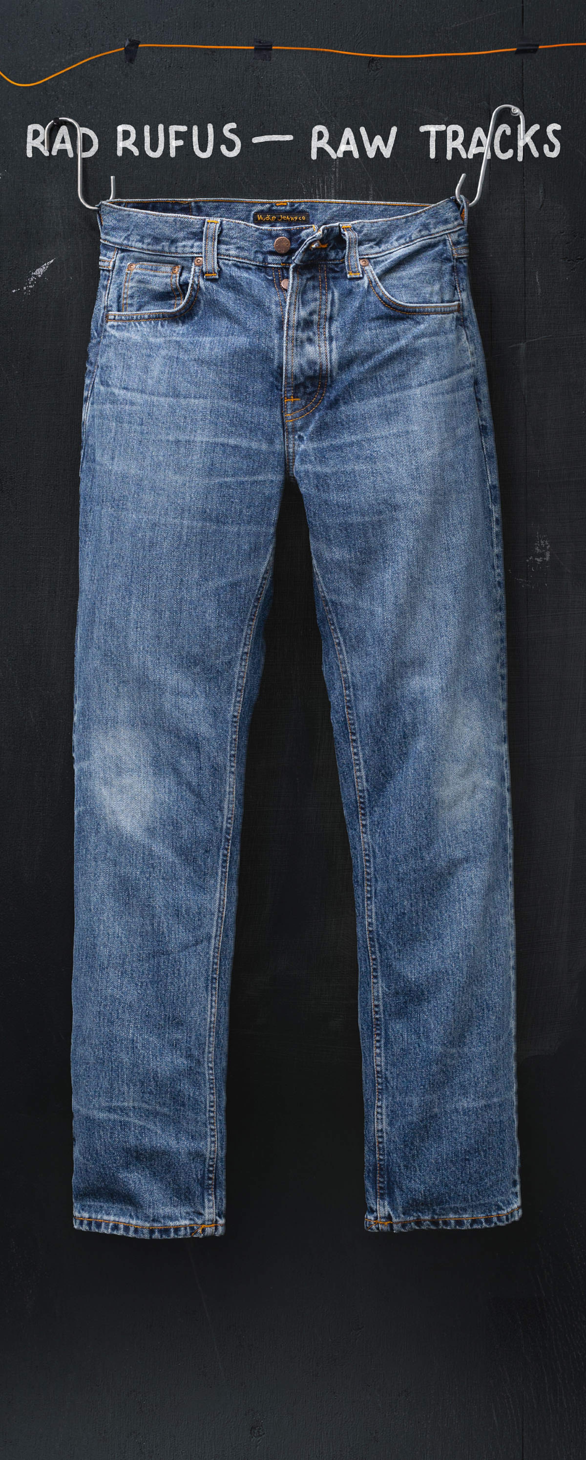 Nudie Jeans Rad Rufus Raw Tracks jeans in faded mid-blue wash on a wire hanger against a chalkboard.