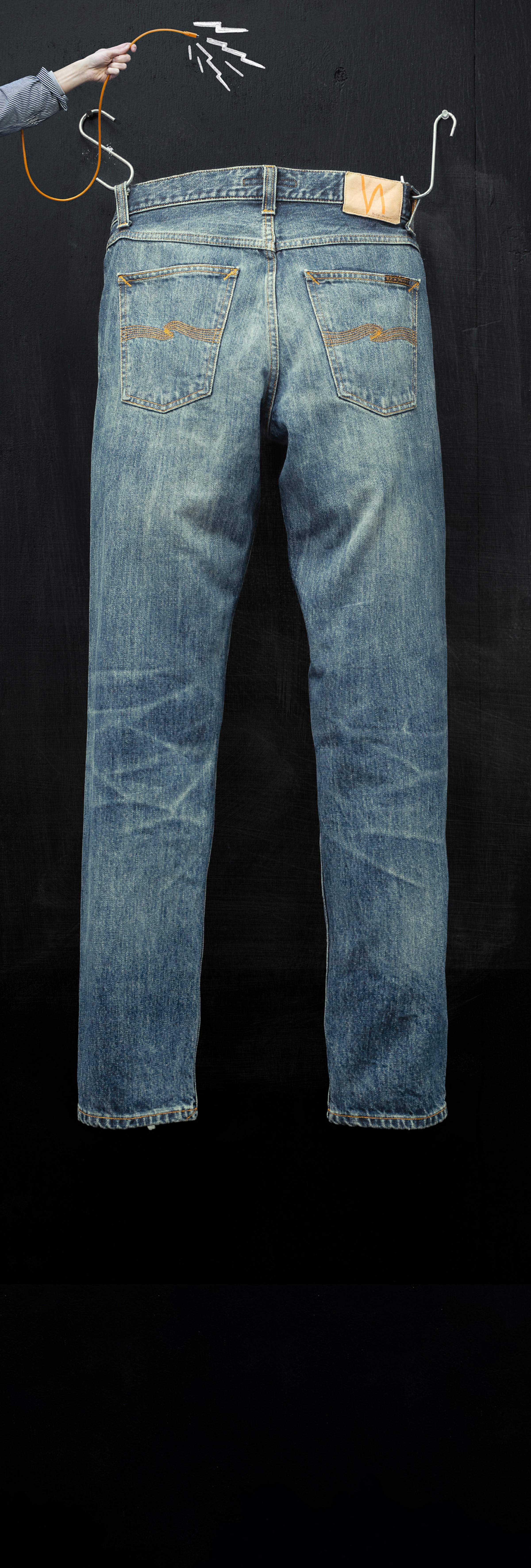 Blue jeans hanging by hangers on a black background, with a hand holding a orange string above.