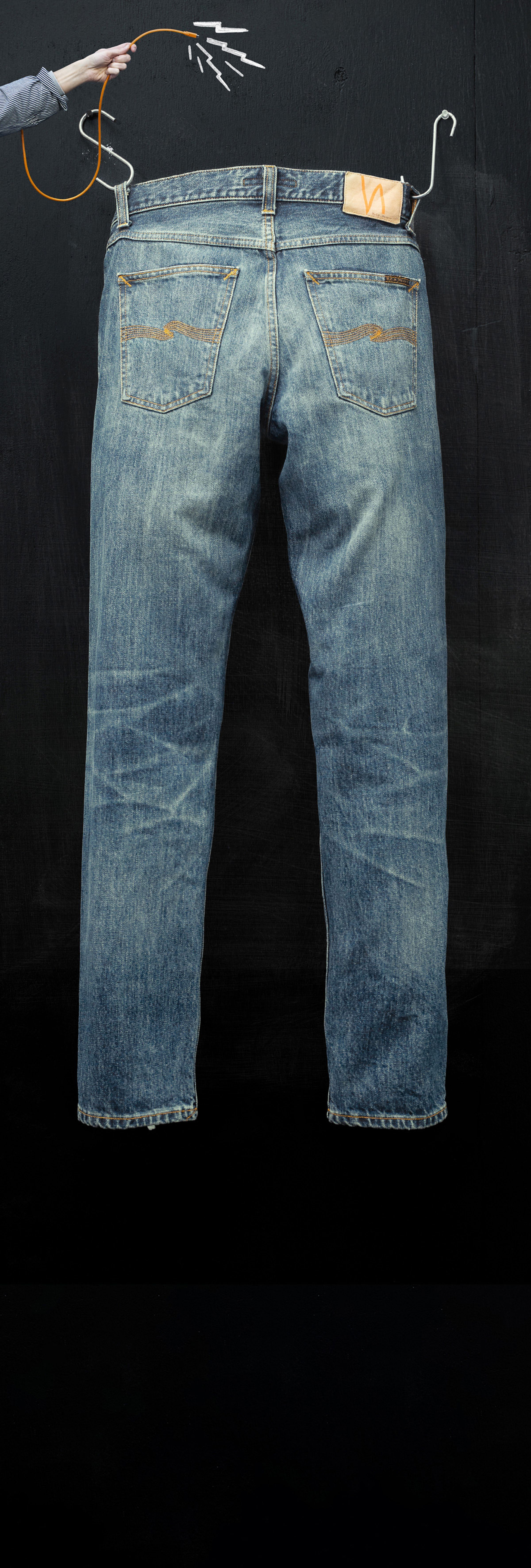 Blue jeans hanging by hangers on a black background, with a hand holding a orange string above.