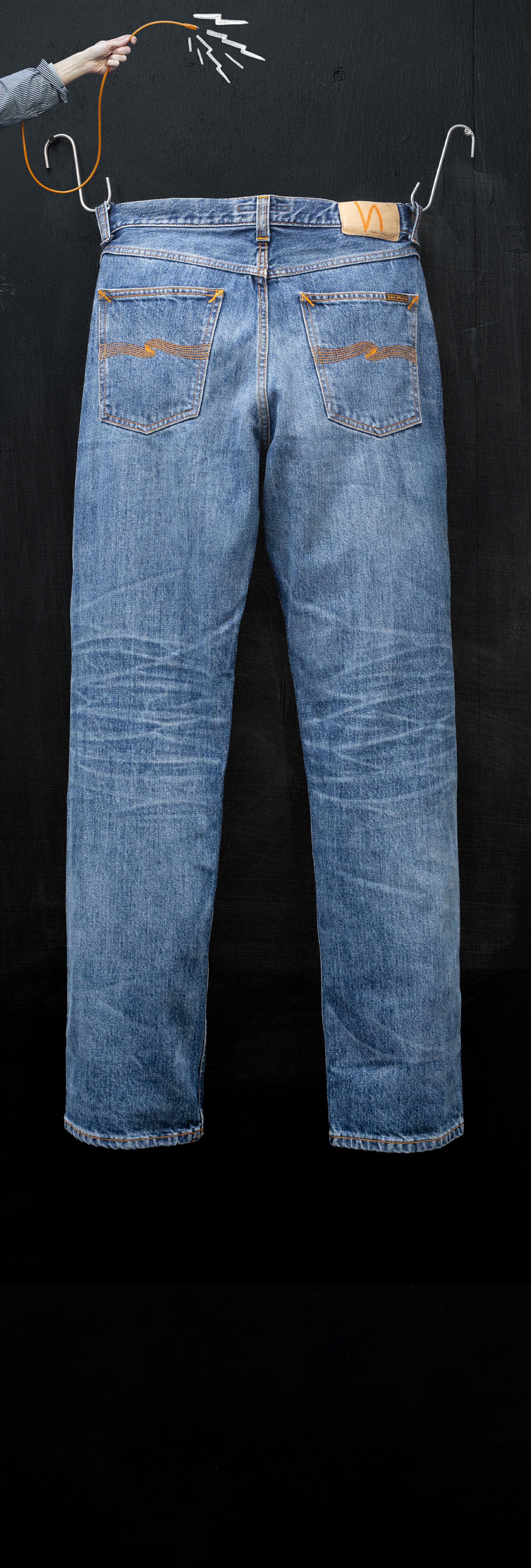 A pair of blue jeans hangs on a black background, suspended by hooks held by a hand.
