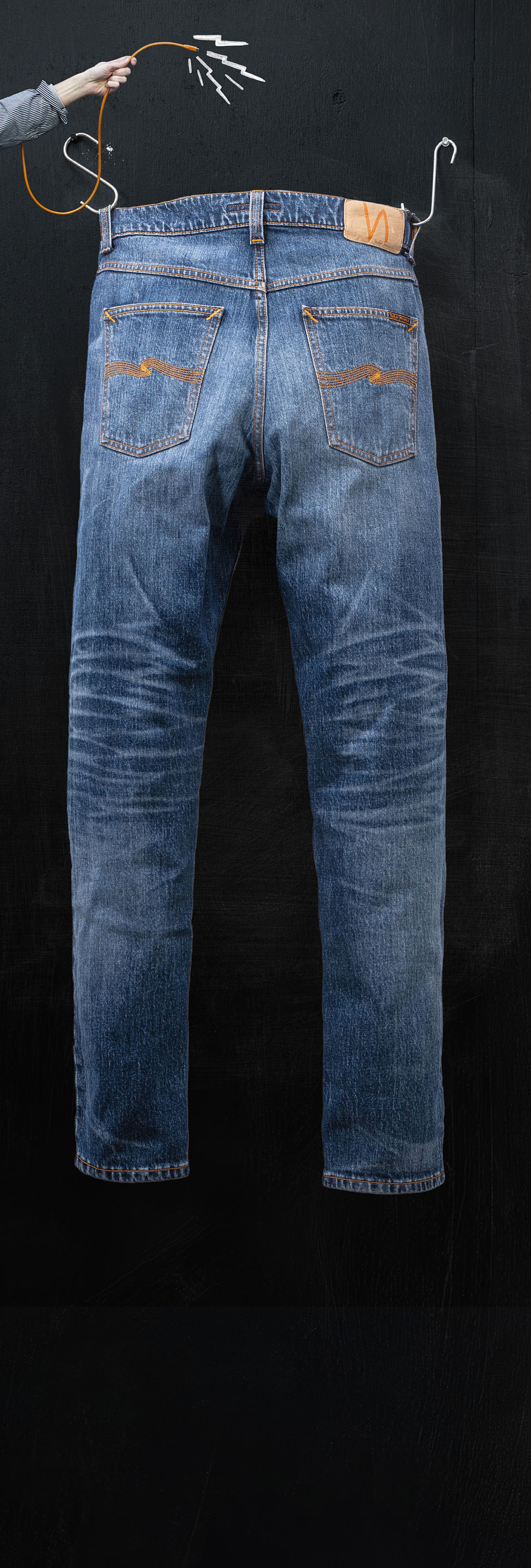 Blue jeans hanging against a dark background, held by a hand using metal hooks.