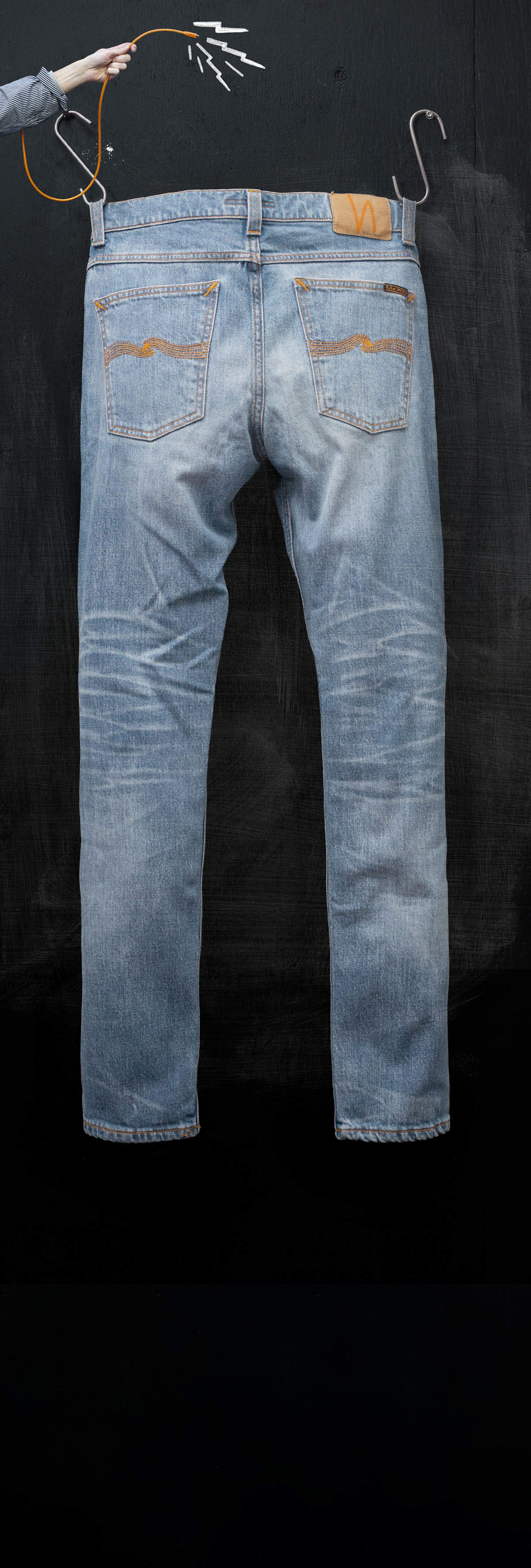 A pair of blue jeans hanging on a black background, held up by a hand with a hanger.