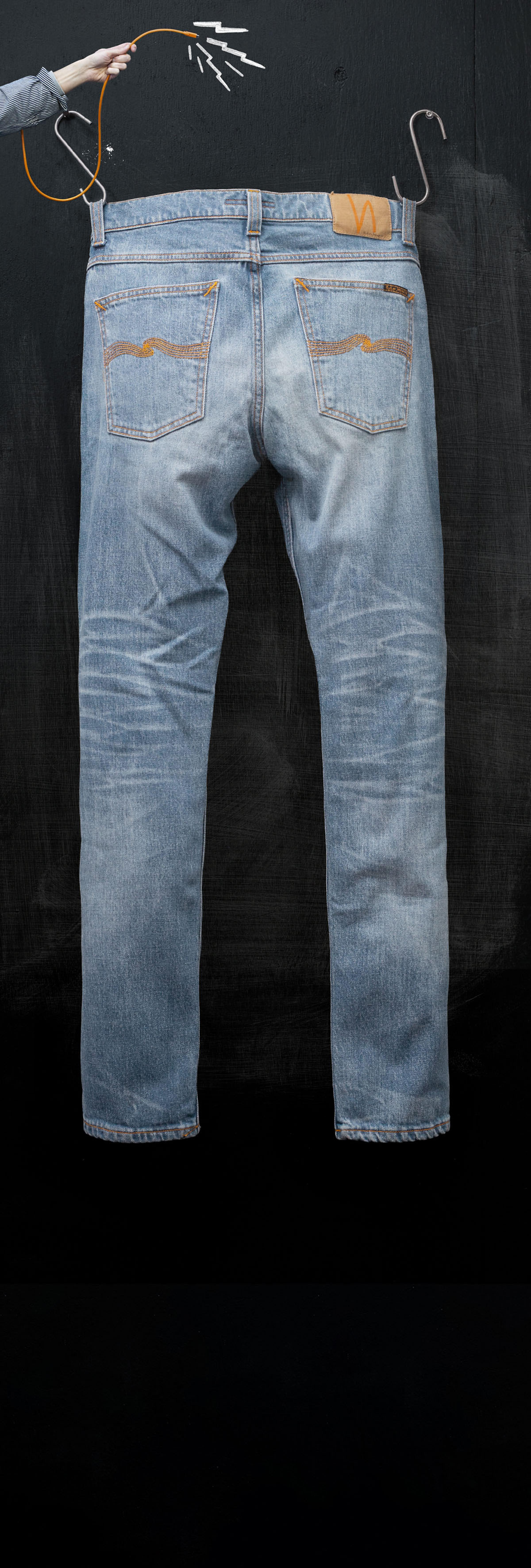 A pair of blue jeans hanging on a black background, held up by a hand with a hanger.