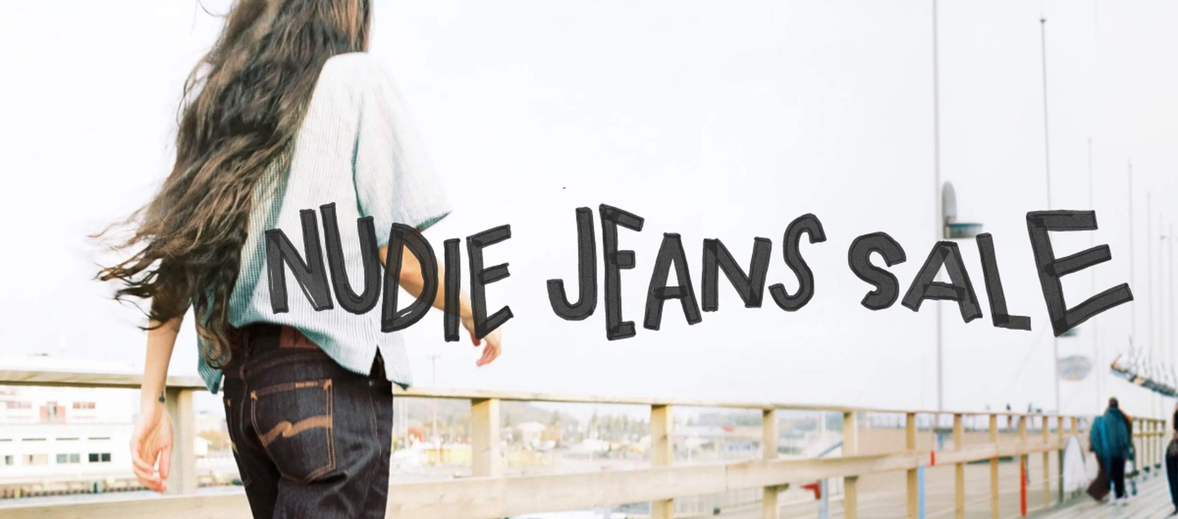 Person in denim and shirt walking on a wooden bridge with urban background, "Nudie Jeans Sale" text overlay.