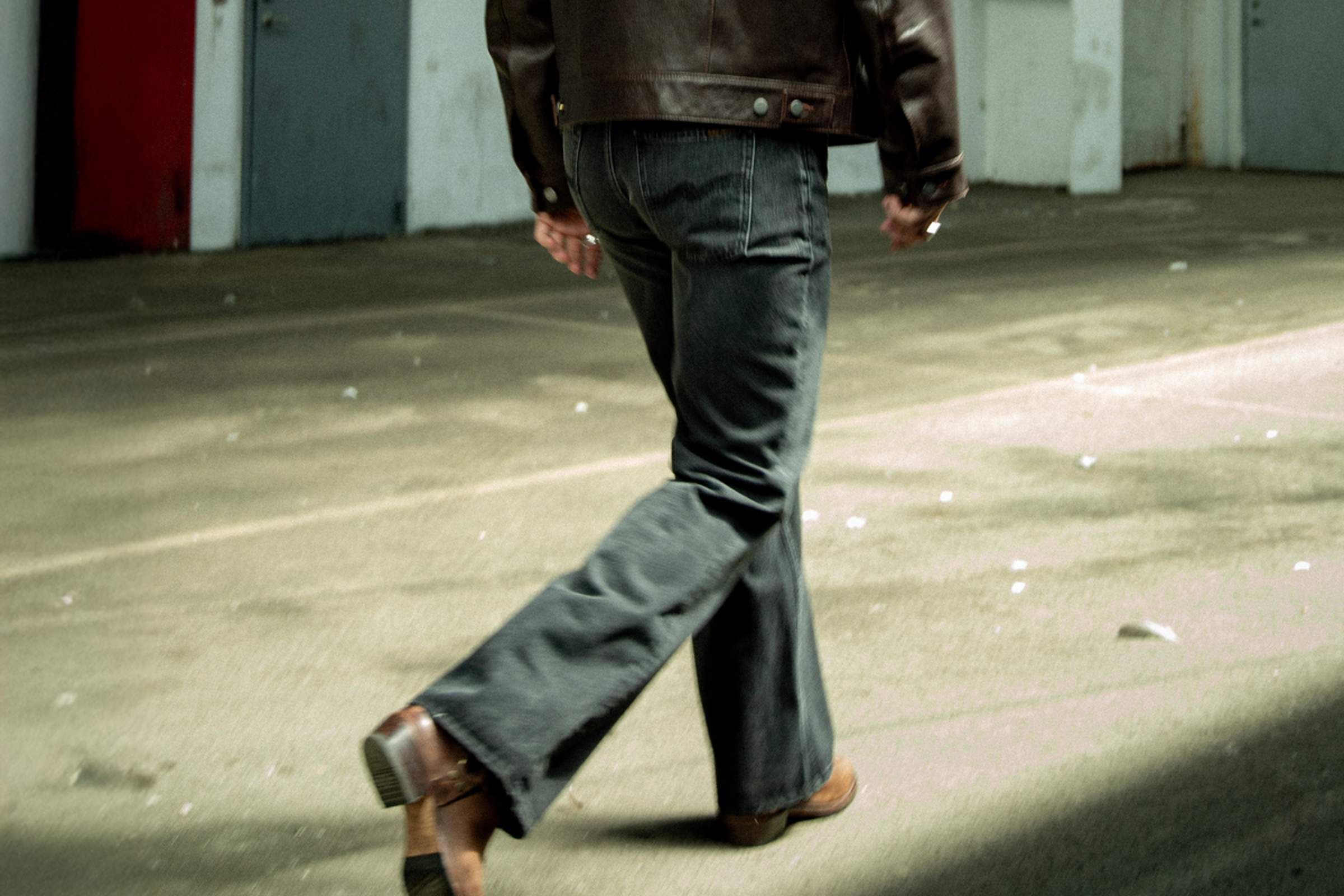 Person in a brown leather jacket and  Nudie Jeans walking away on a sunlit concrete surface, casting a shadow.