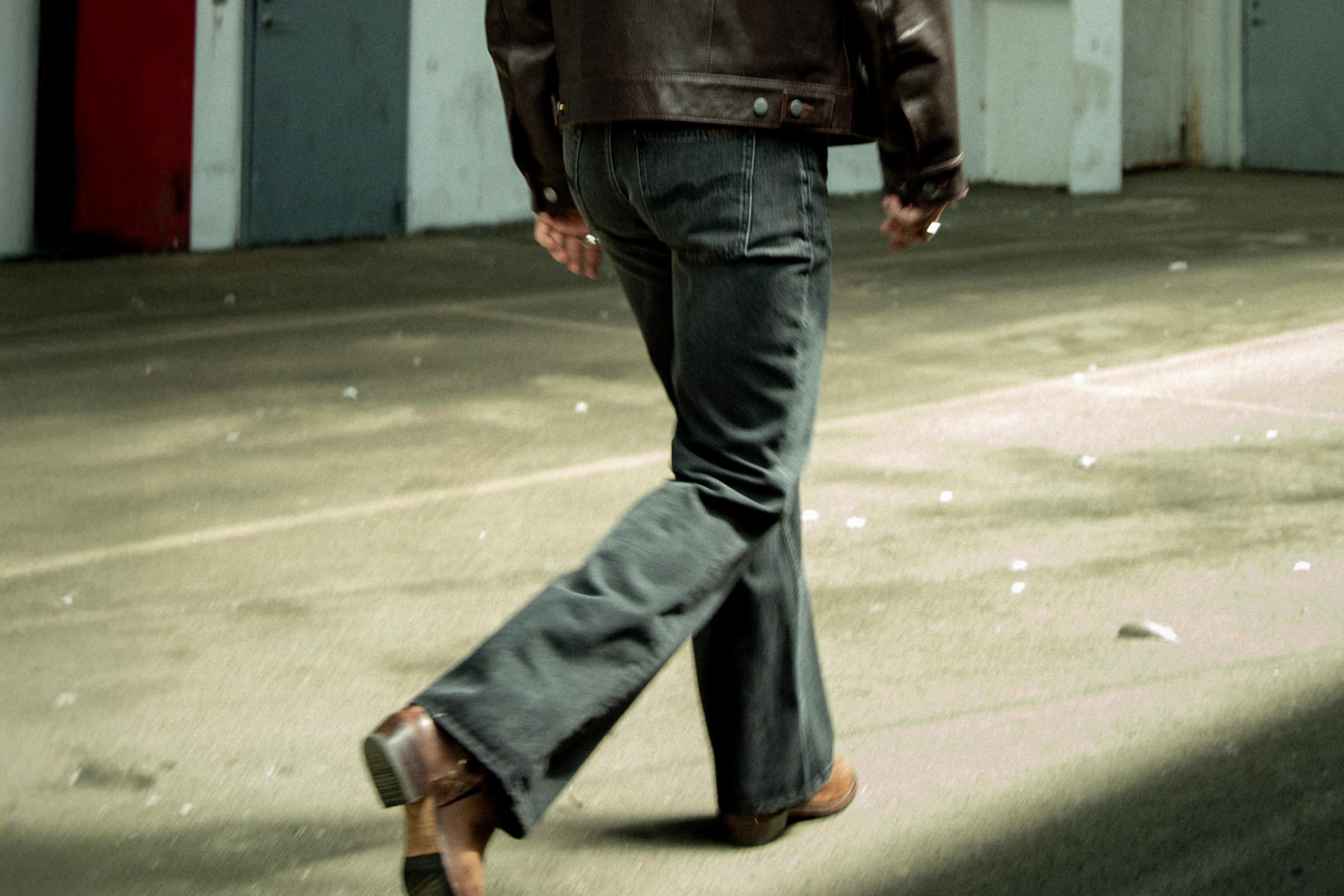 Person in a brown leather jacket and  Nudie Jeans walking away on a sunlit concrete surface, casting a shadow.