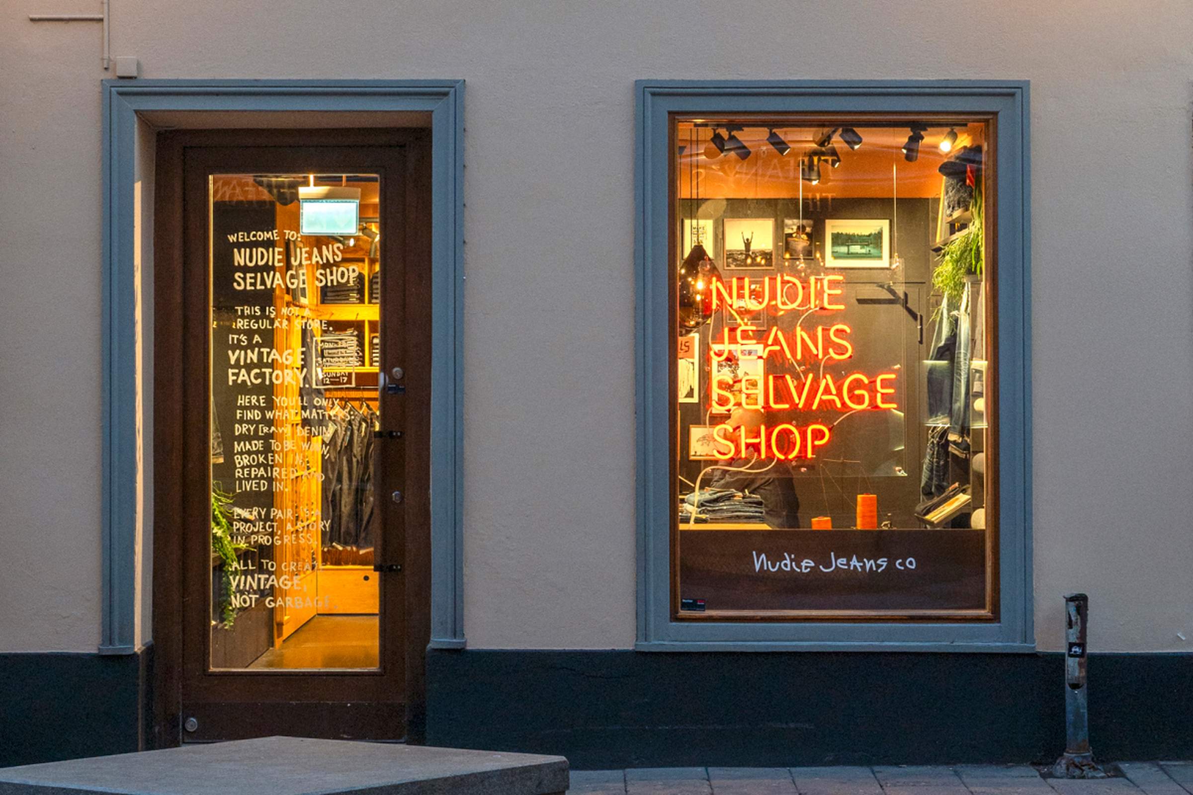 Storefront with two windows displaying "Nudie Jeans Selvage Shop" in neon. One window shows apparel; the door has text and reflections.