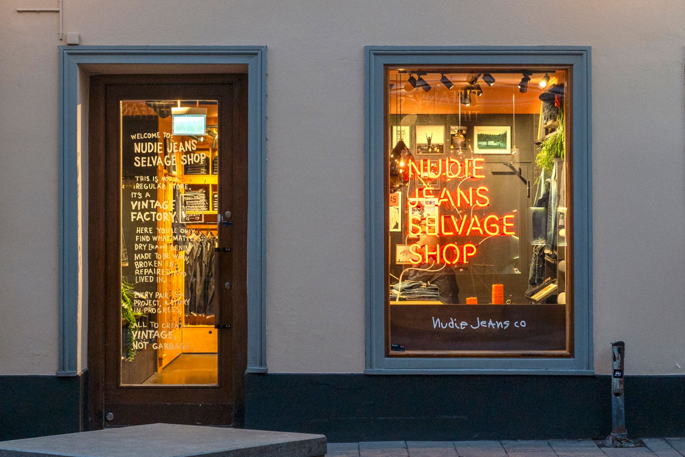 Storefront with two windows displaying "Nudie Jeans Selvage Shop" in neon. One window shows apparel; the door has text and reflections.