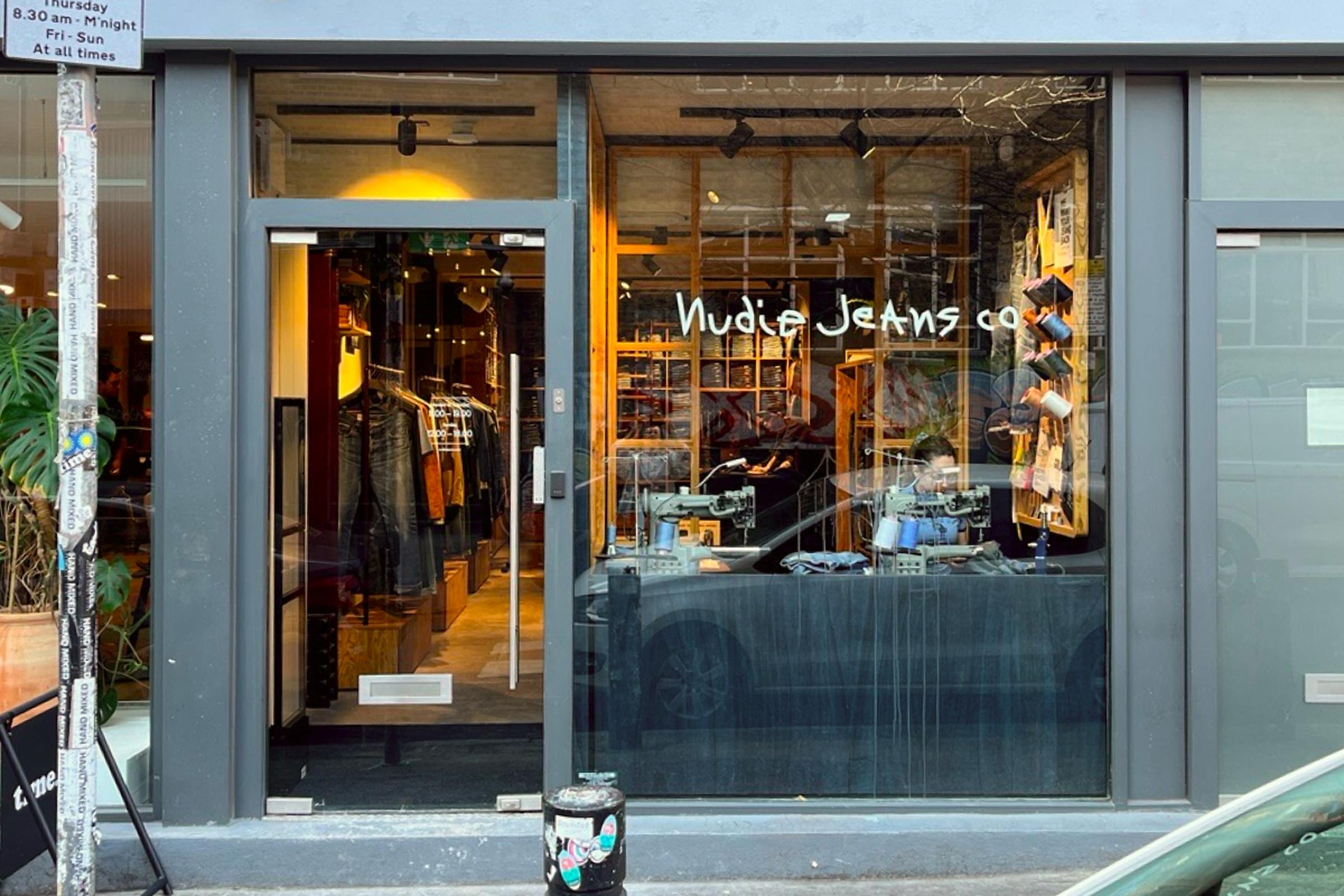 Storefront of Nudie Jeans Co. with large glass windows displaying clothing racks inside, and a small plant and sign outside.