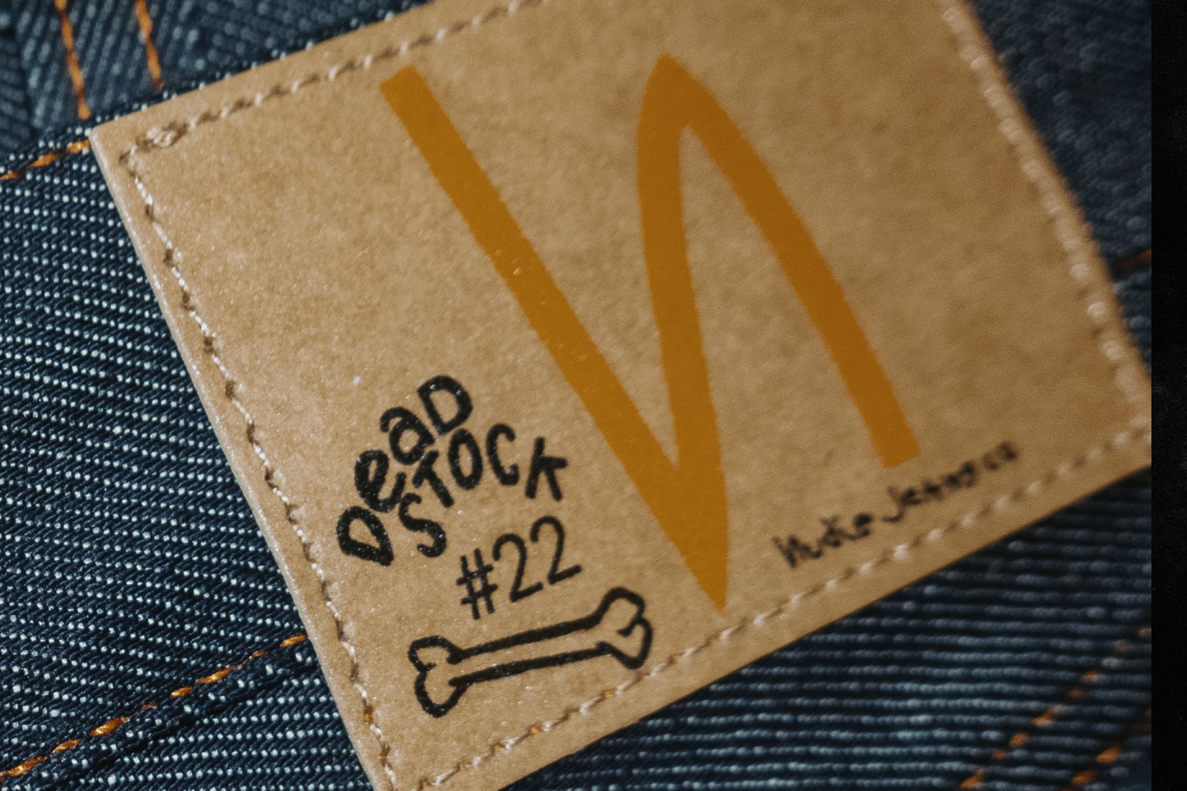 Close-up of a denim fabric with a brown label featuring text "Dead Stock #22" and a drawing of a bone, alongside a large orange "N".