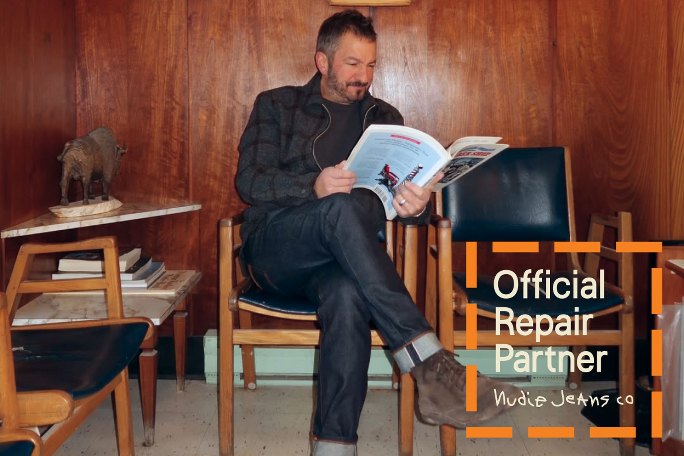 Man in a cozy room, seated and reading a magazine. Text overlay: "Official Repair Partner Nudie Jeans Co."