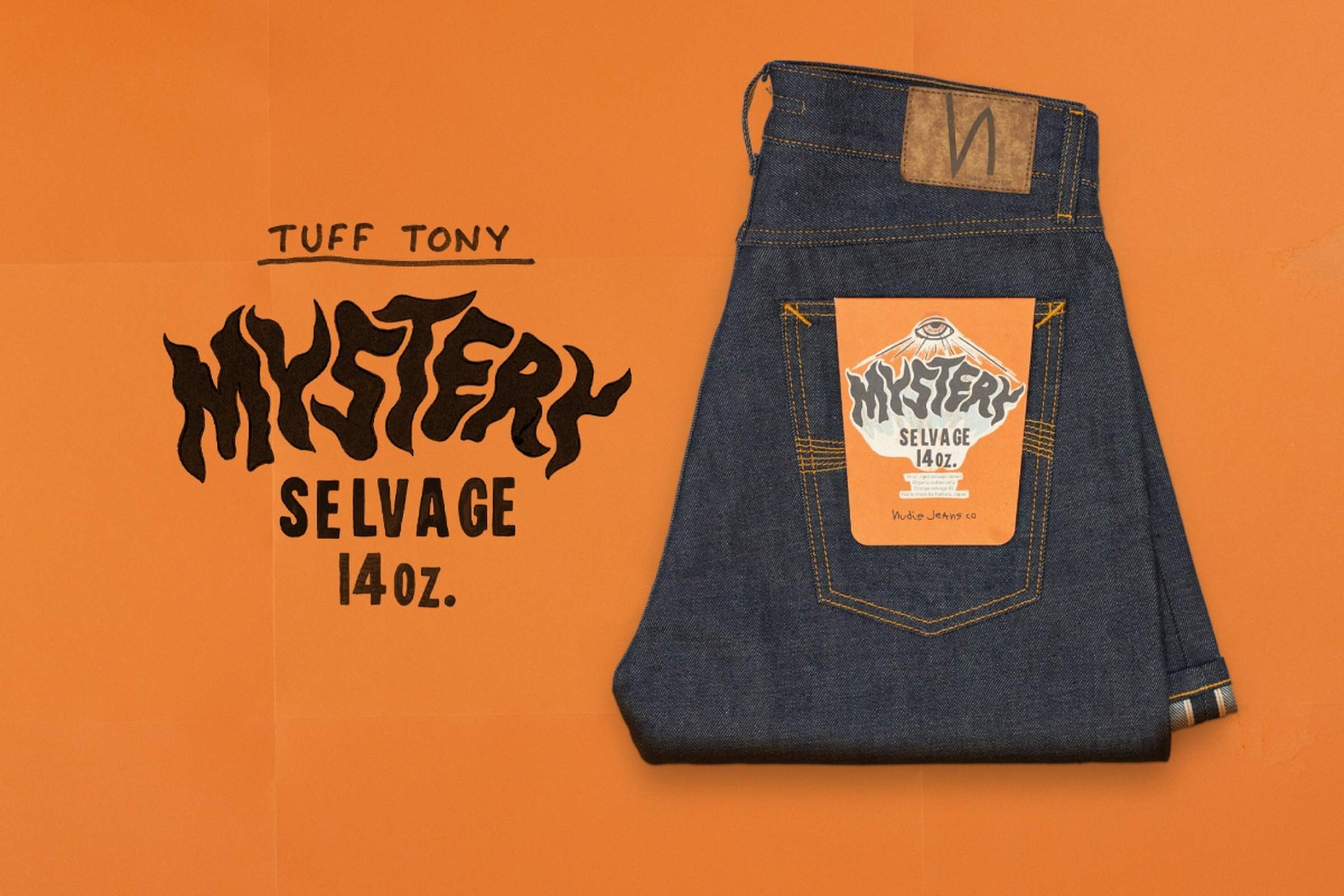 Folded pair of dark blue jeans with a colorful label on the back pocket, placed on an orange background. Mystery Selvage. Nudie Jeans