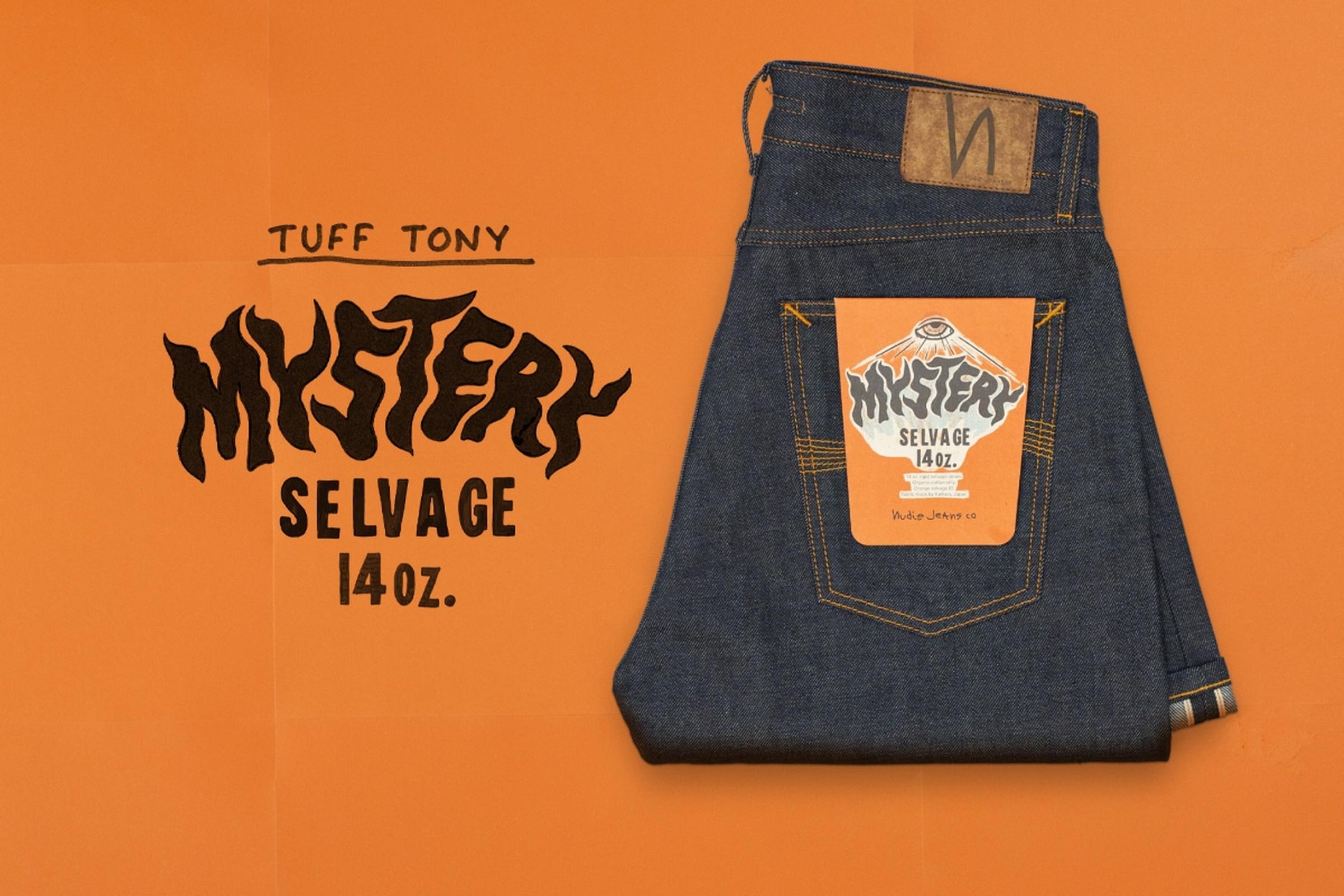 Folded pair of dark blue jeans with a colorful label on the back pocket, placed on an orange background. Mystery Selvage. Nudie Jeans