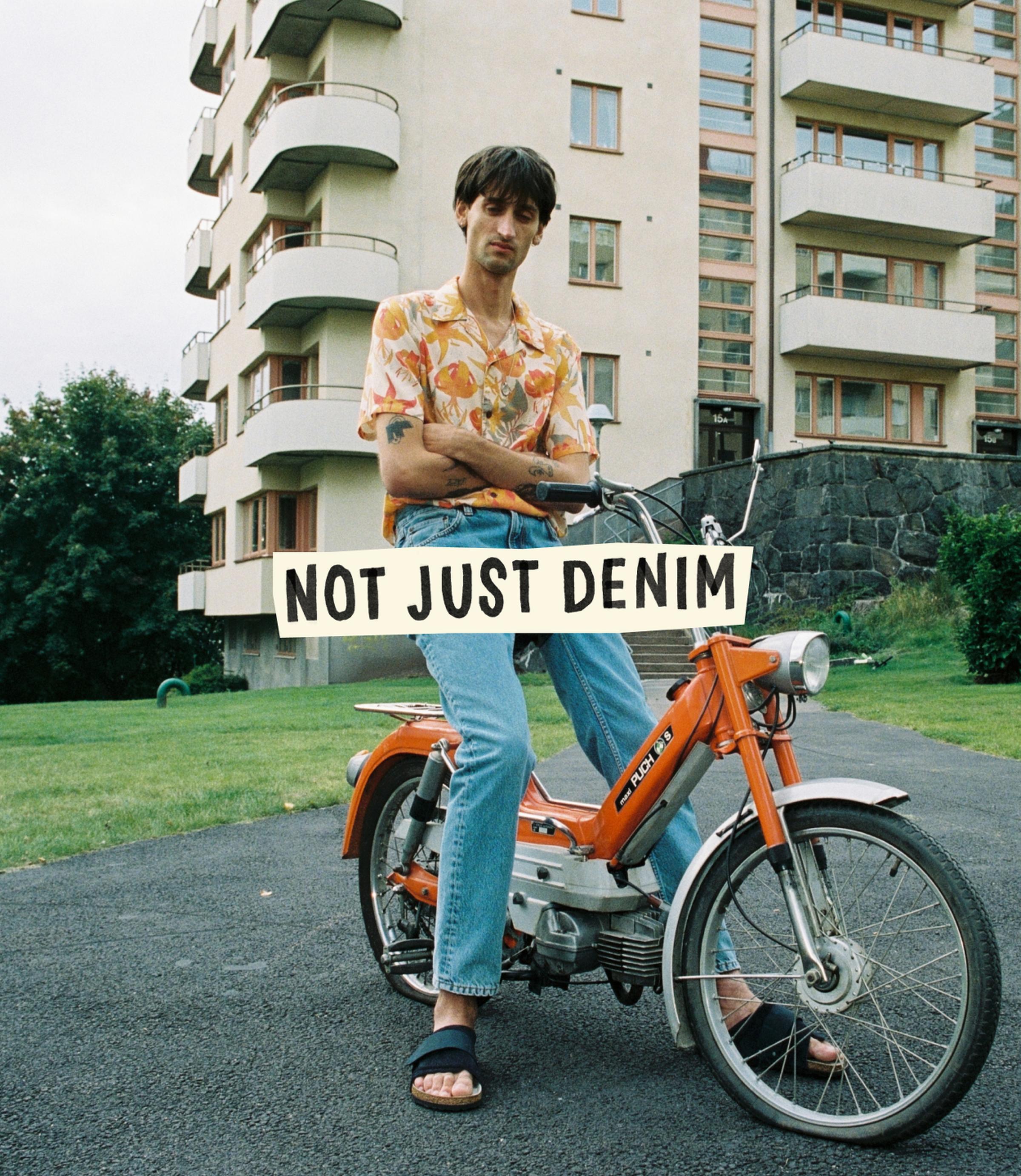 Man in a floral shirt and jeans sits on an orange moped in front of an apartment building, with a "Not Just Denim" sign overlay.