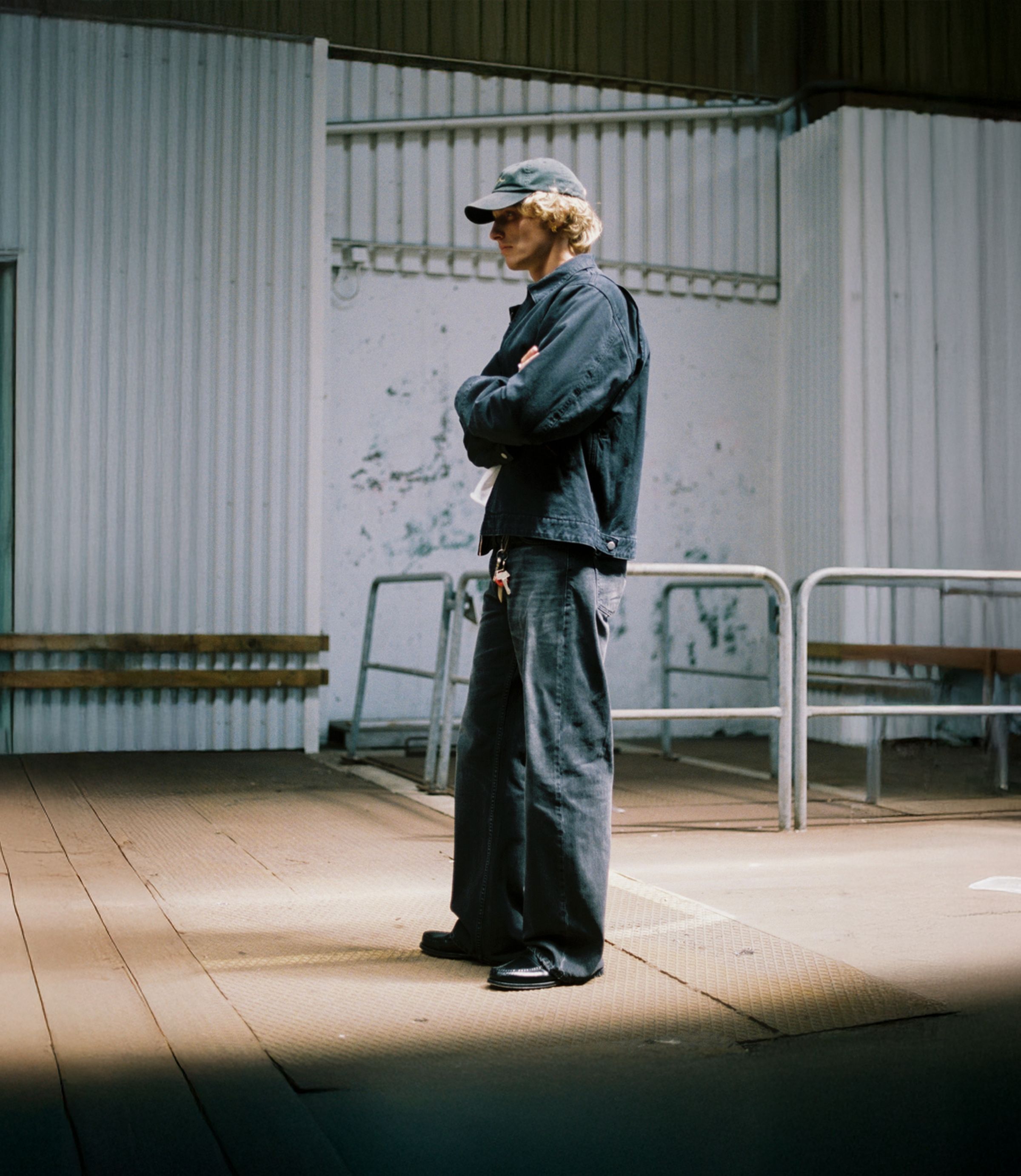 Person in dark clothing and cap stands with arms crossed in an industrial setting, with corrugated metal walls and a partially visible word.