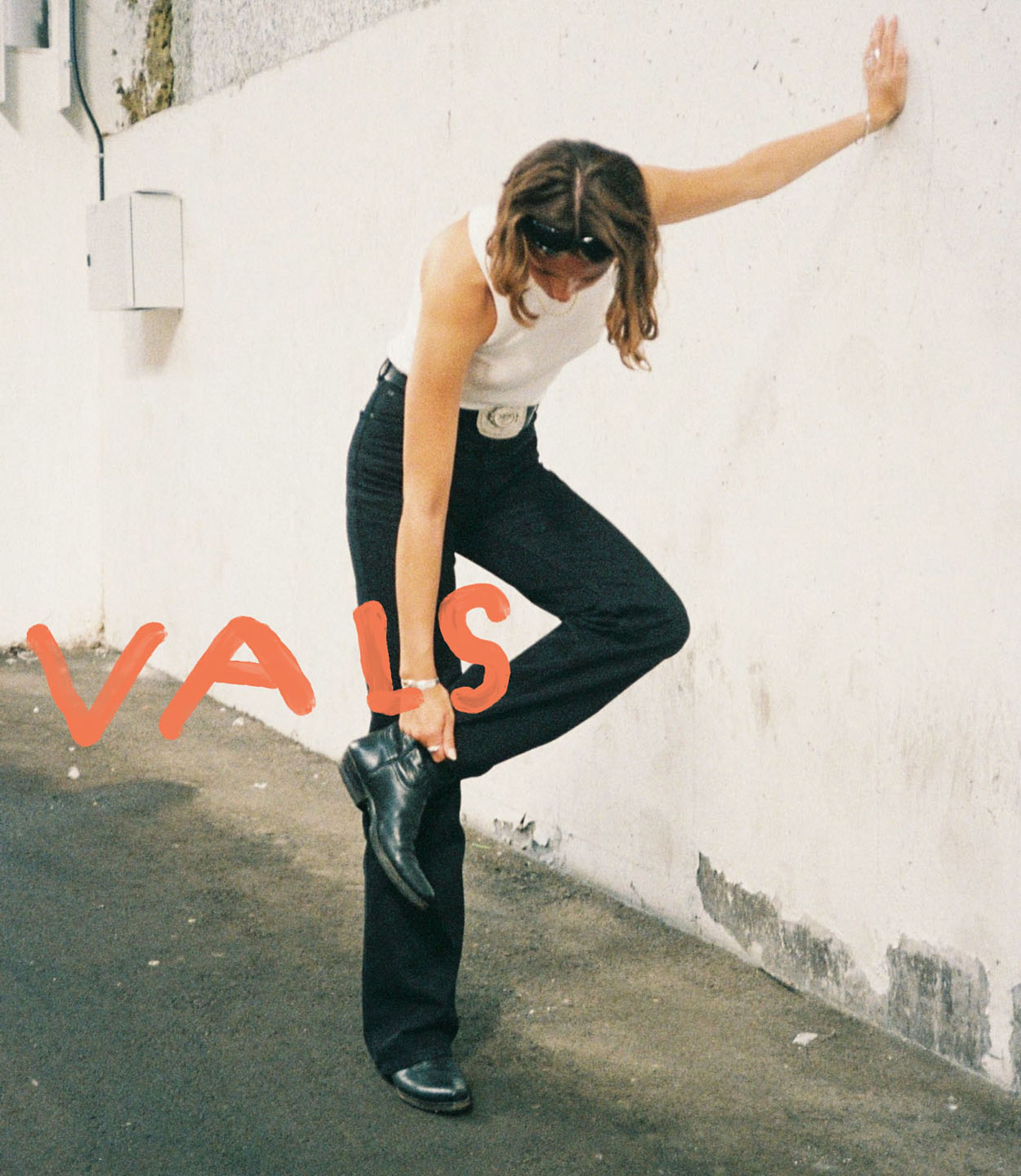 Person in a white tank top and black pants adjusts their boot against a white wall. Red text reads "VAIS."