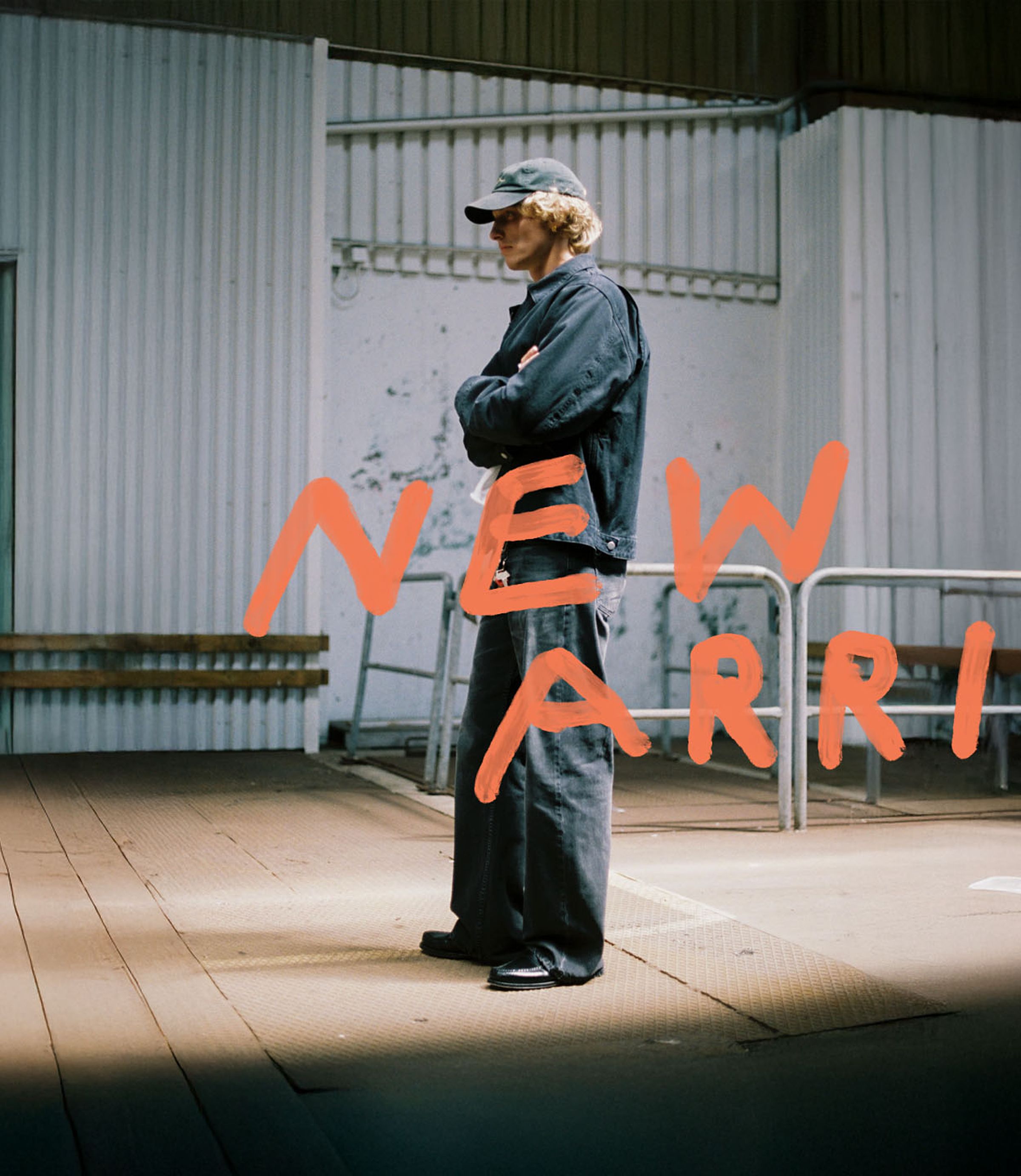 Person in dark clothing and cap stands with arms crossed in an industrial space. The words "NEW ARRIVAL" are written in orange across the image.