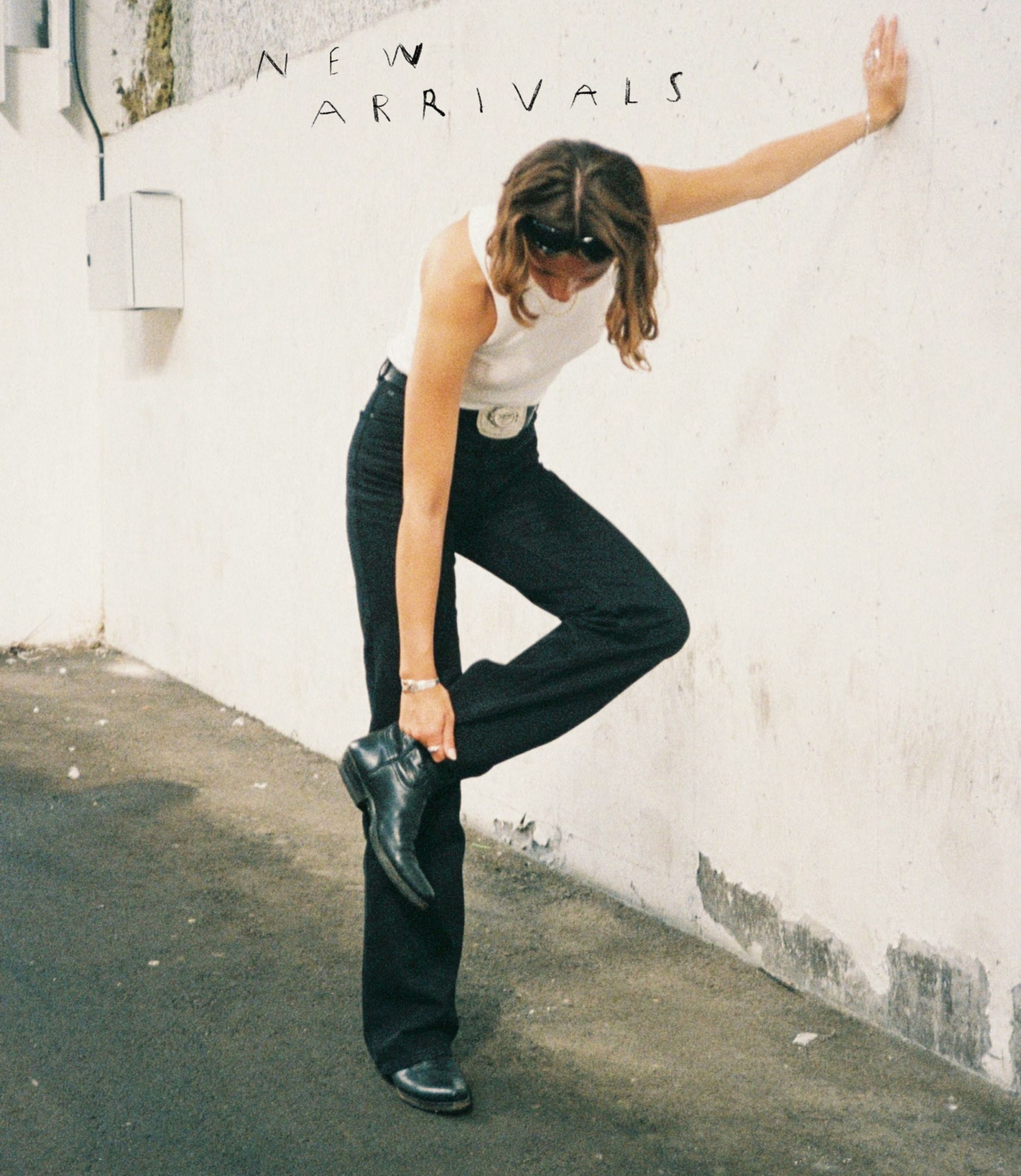 Person in a white tank top and black pants leans against a wall adjusting a boot, with "NEW ARR" text on the wall.