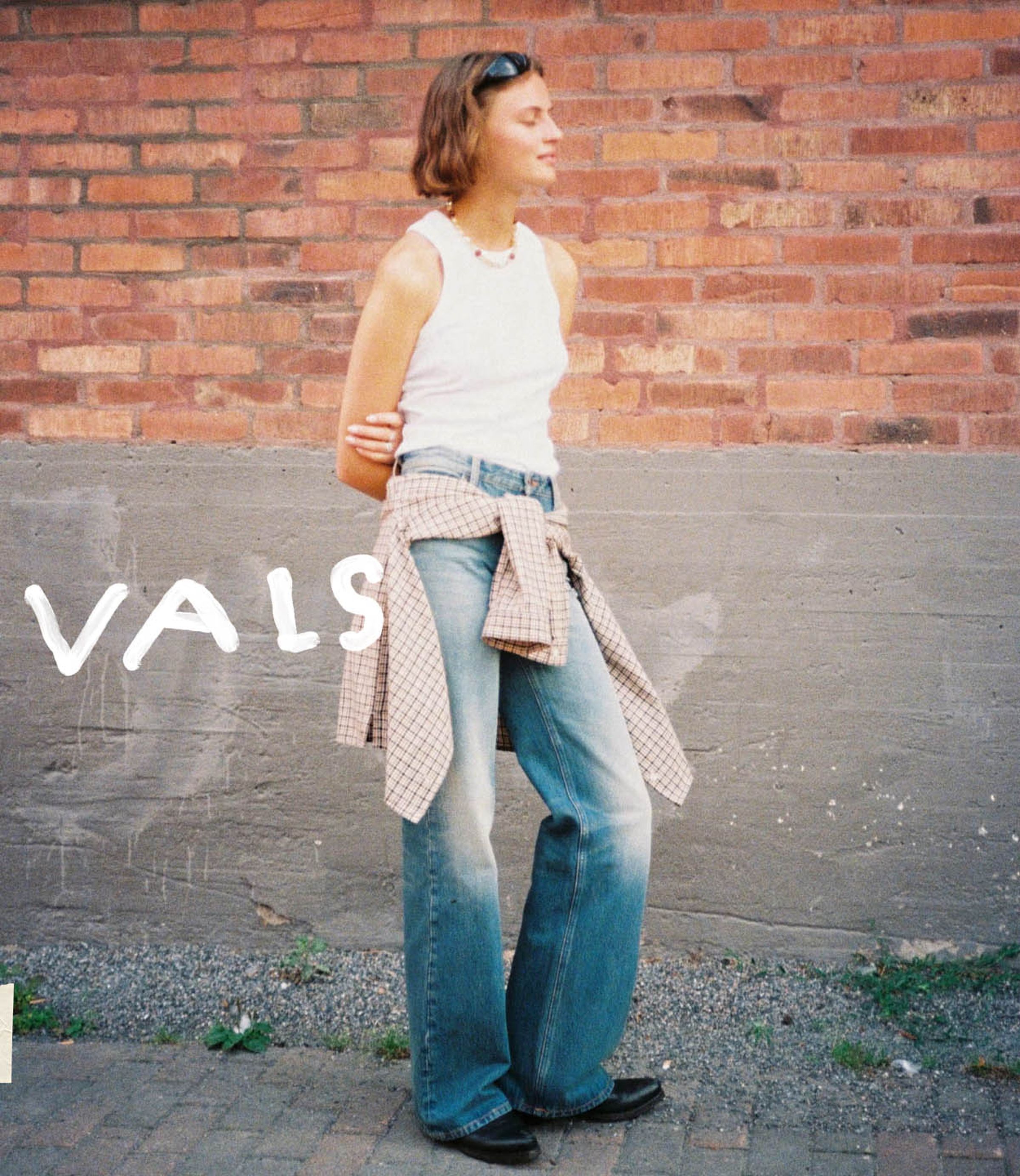 Person in a white tank top and jeans stands against a brick wall, with a plaid shirt tied at the waist.