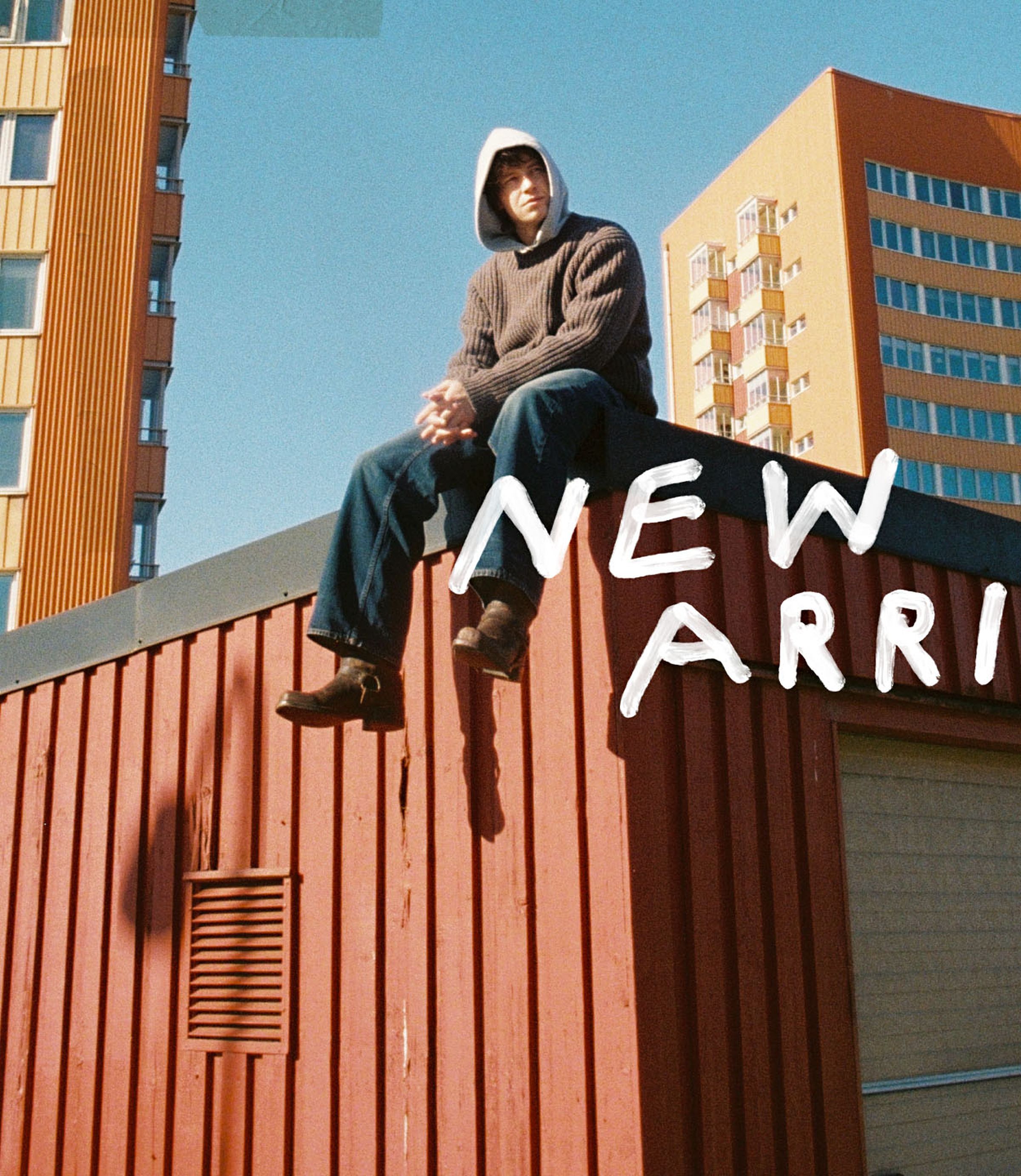 Person in a hoodie and and Nuide Jeans sits on a red rooftop with modern buildings in the background. Text reads "New Arrivals."