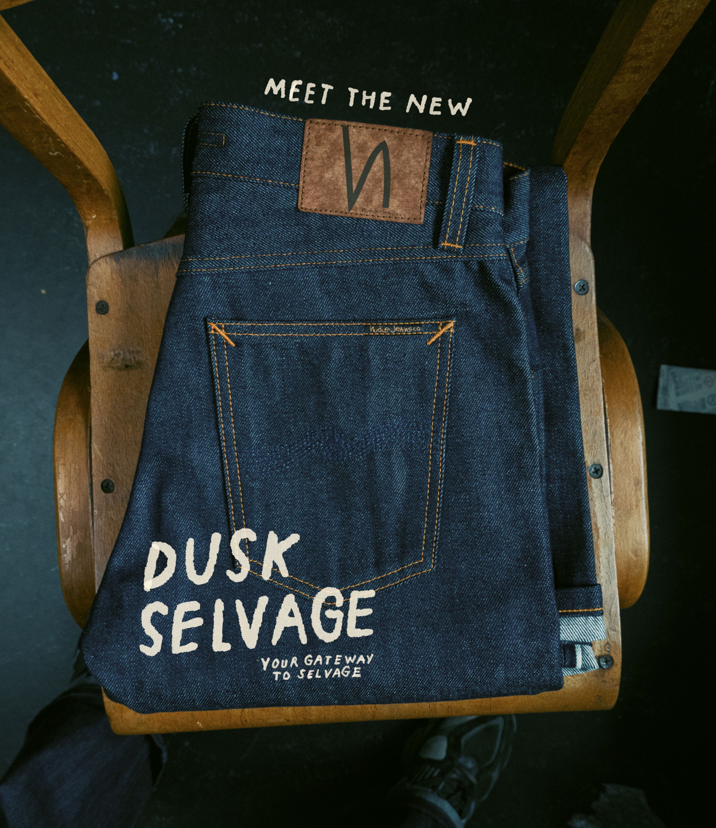 RAW selvage denim jeans with brown leather patch on a wooden chair. Text reads, "Meet the New Dusk Selvage: Your Gateway to Selvage."