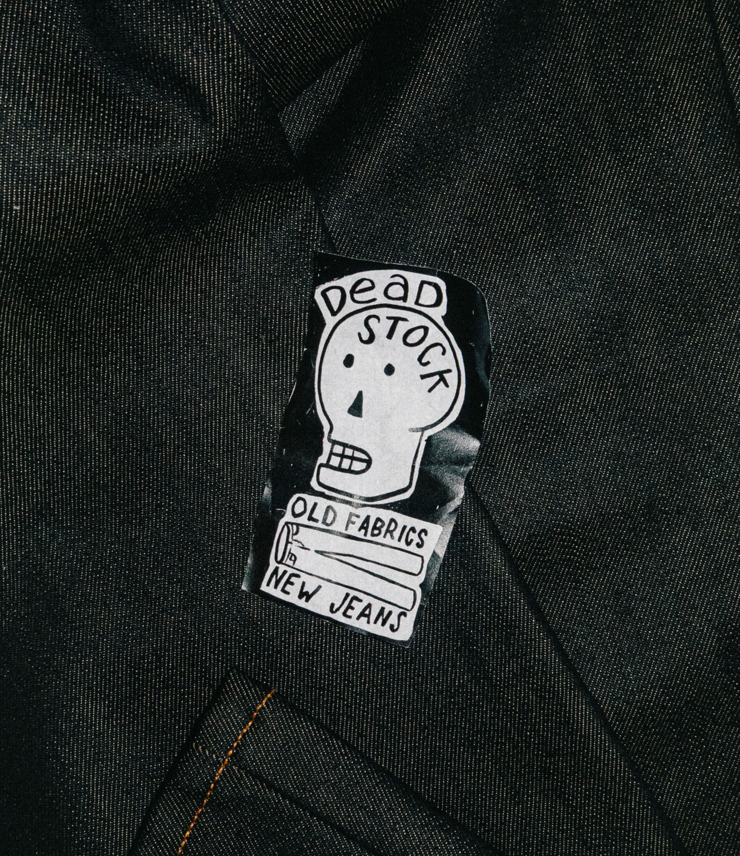 A black fabric patch with a white skull illustration and the text "Dead Stock, Old Fabrics, New Jeans" sewn onto dark denim.