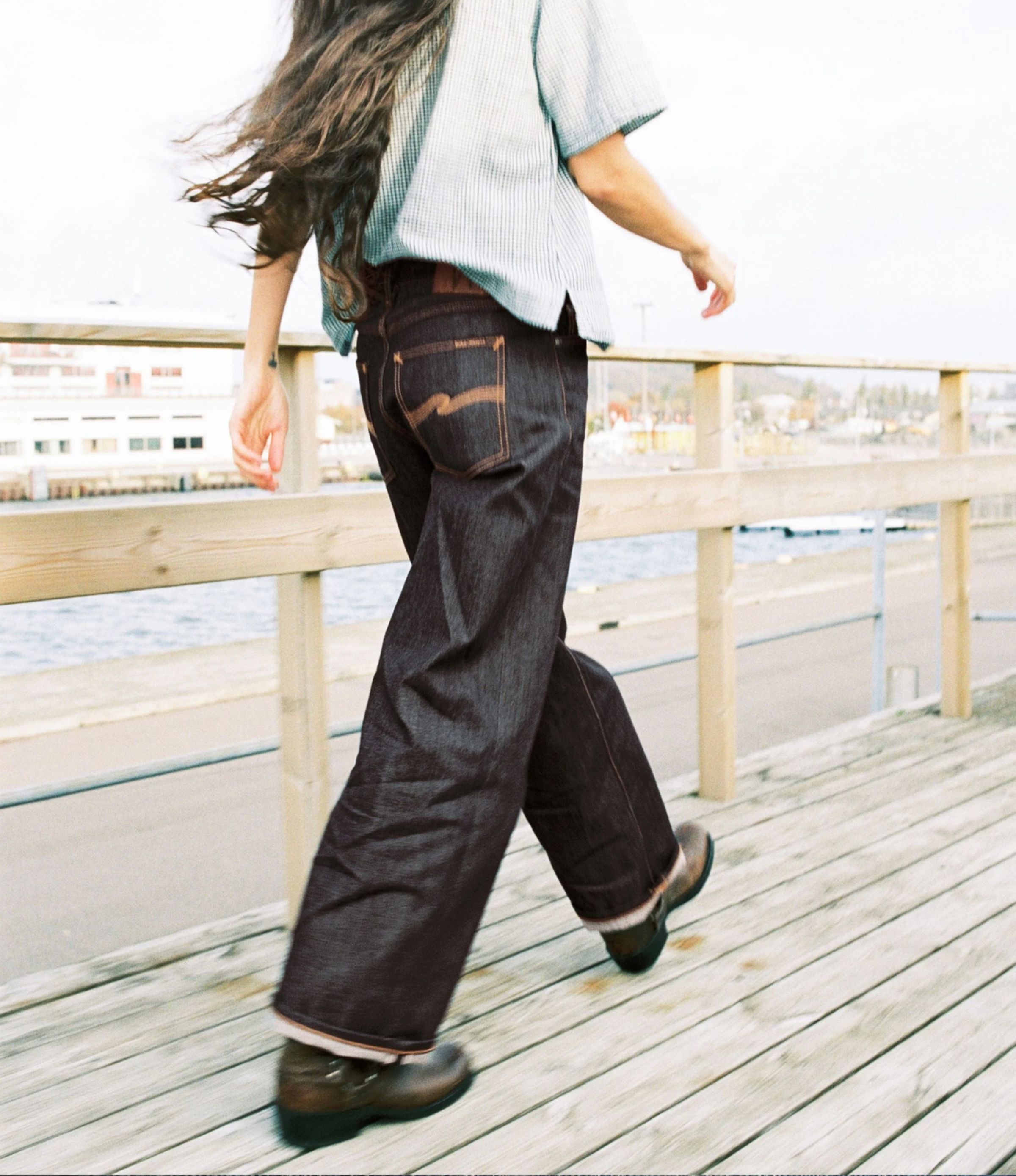 Person walking on a wooden boardwalk by the water, wearing a striped shirt and wide-leg jeans, with long hair flowing behind.