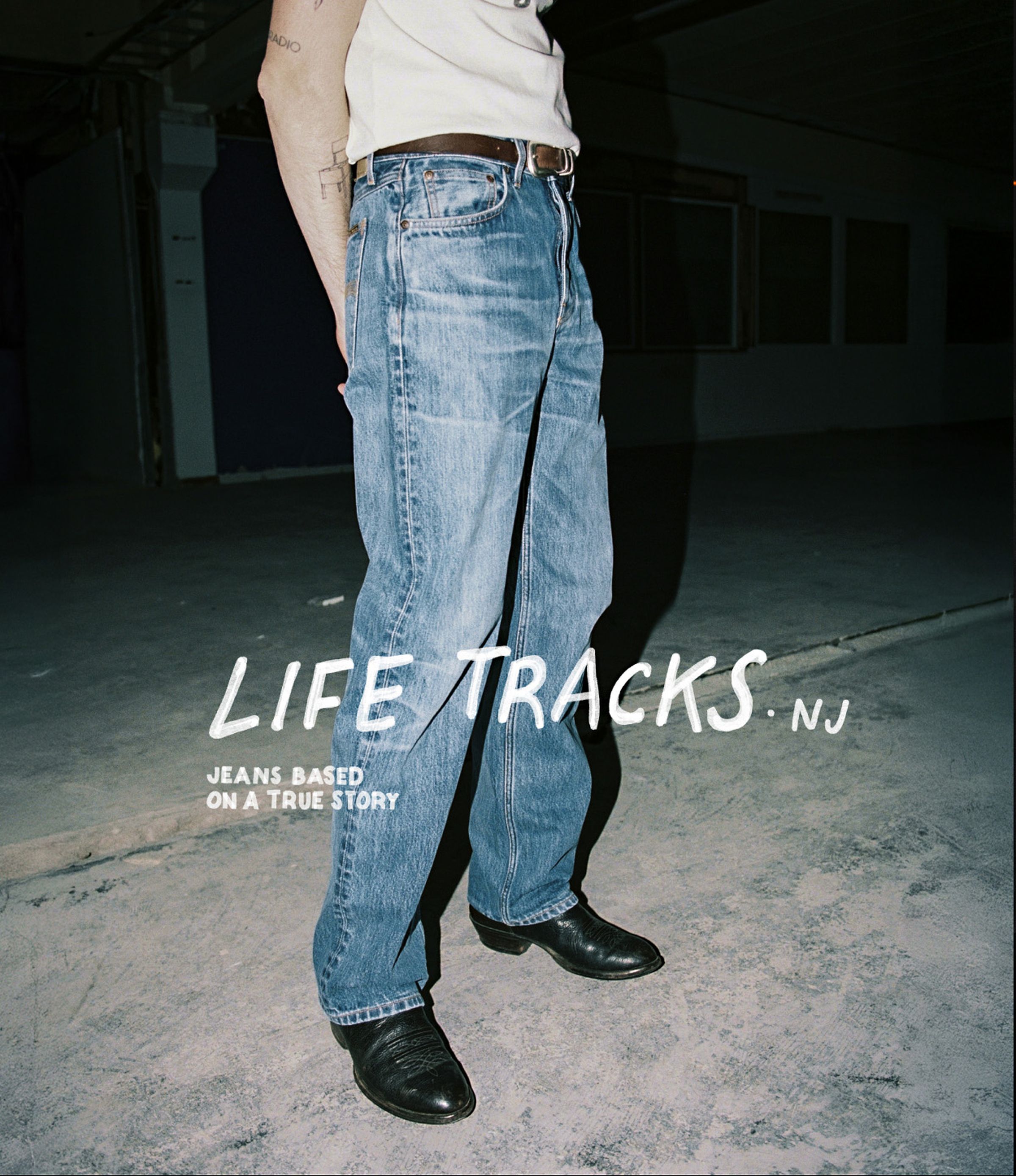 Person wearing blue jeans and black boots stands indoors on a concrete floor. Text on the image reads "LIFE TRACKS NJ" and "JEANS BASED ON A TRUE STORY."