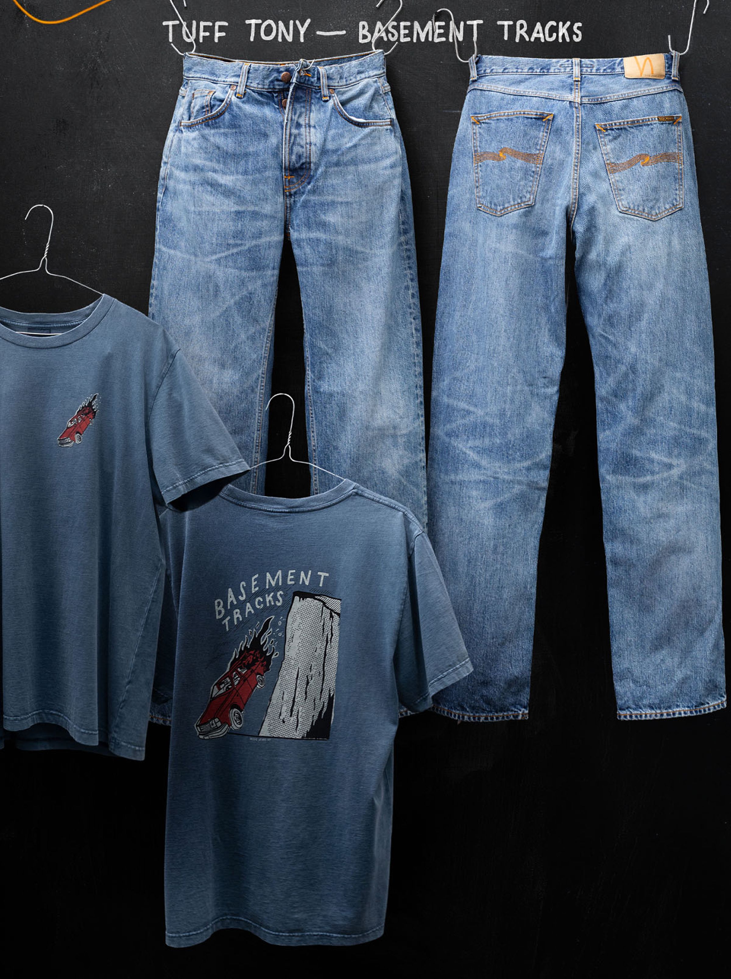 Two pairs of blue jeans and two blue graphic T-shirts with "Basement Tracks" designs hang against a black background.