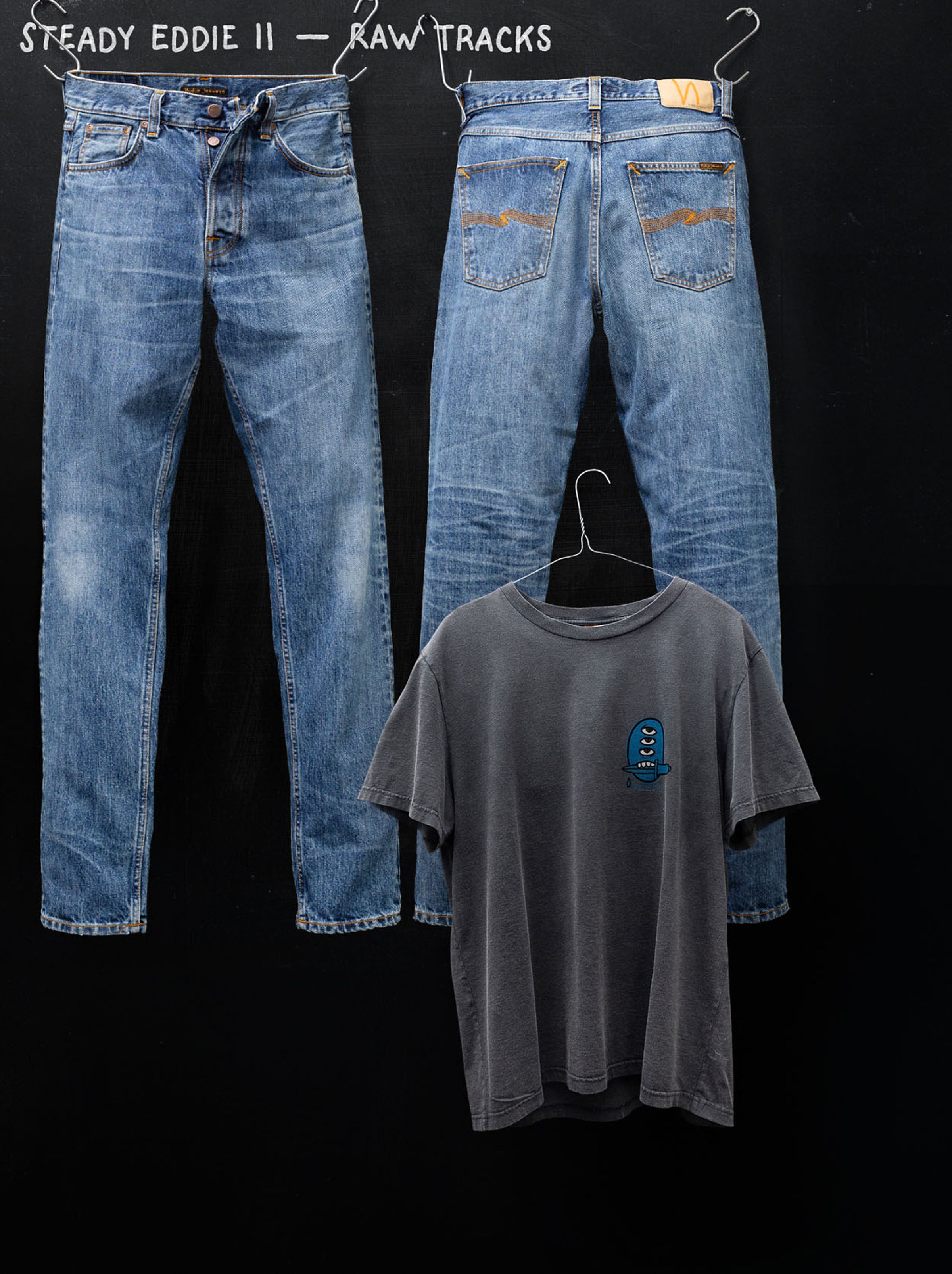 Two pairs of blue jeans and a black T-shirt with a small blue graphic hang against a dark background.