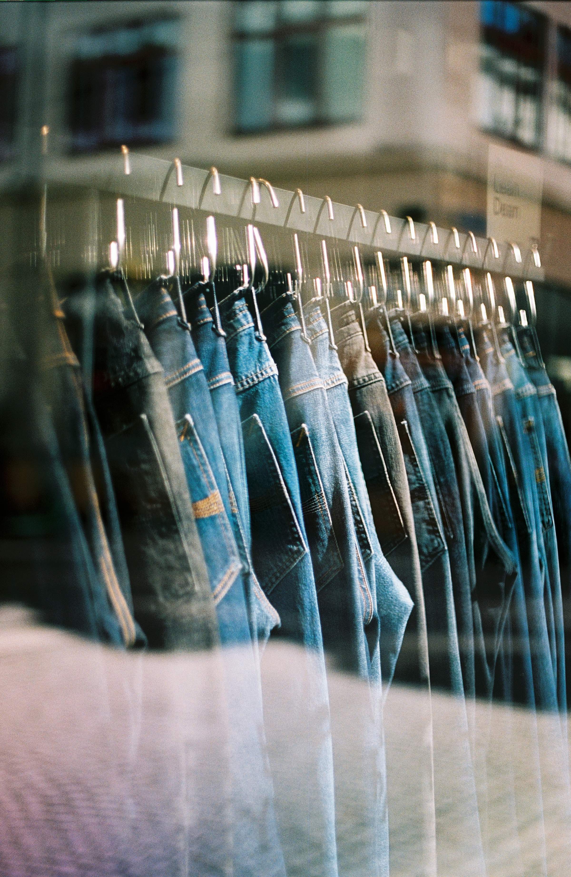 A row of Nudie Jeans hanging on a rack in a store, reflected in a glass window with a blurred street view in the background.